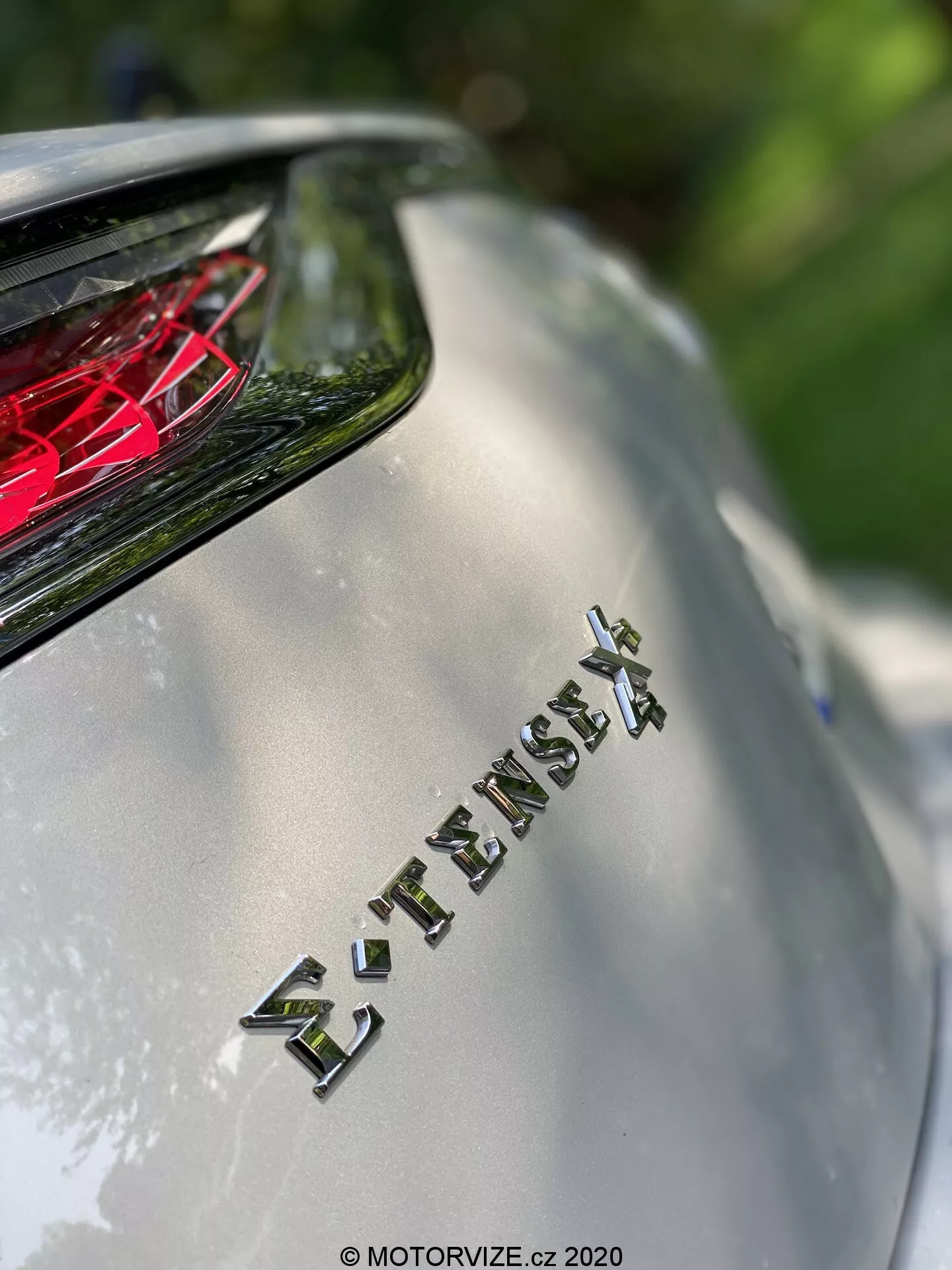Pogled od blizu na zadnji del modela DS Automobiles DS 7 Crossback 2018, s poudarkom na emblemu 'E-TENSE 4X4', ki kaže na štirikolesni pogon in verjetno hibridni ali električni pogon. Majhna globinska ostrina zamegli ozadje, medtem ko poudari emblem, nad katerim je vidna rdeča kristalna zadnja luč. Slika zajame kovinsko površino karoserije in kromirane črke v naravni sončni svetlobi, kar poudarja napredno tehnologijo in luksuzno obliko vozila.