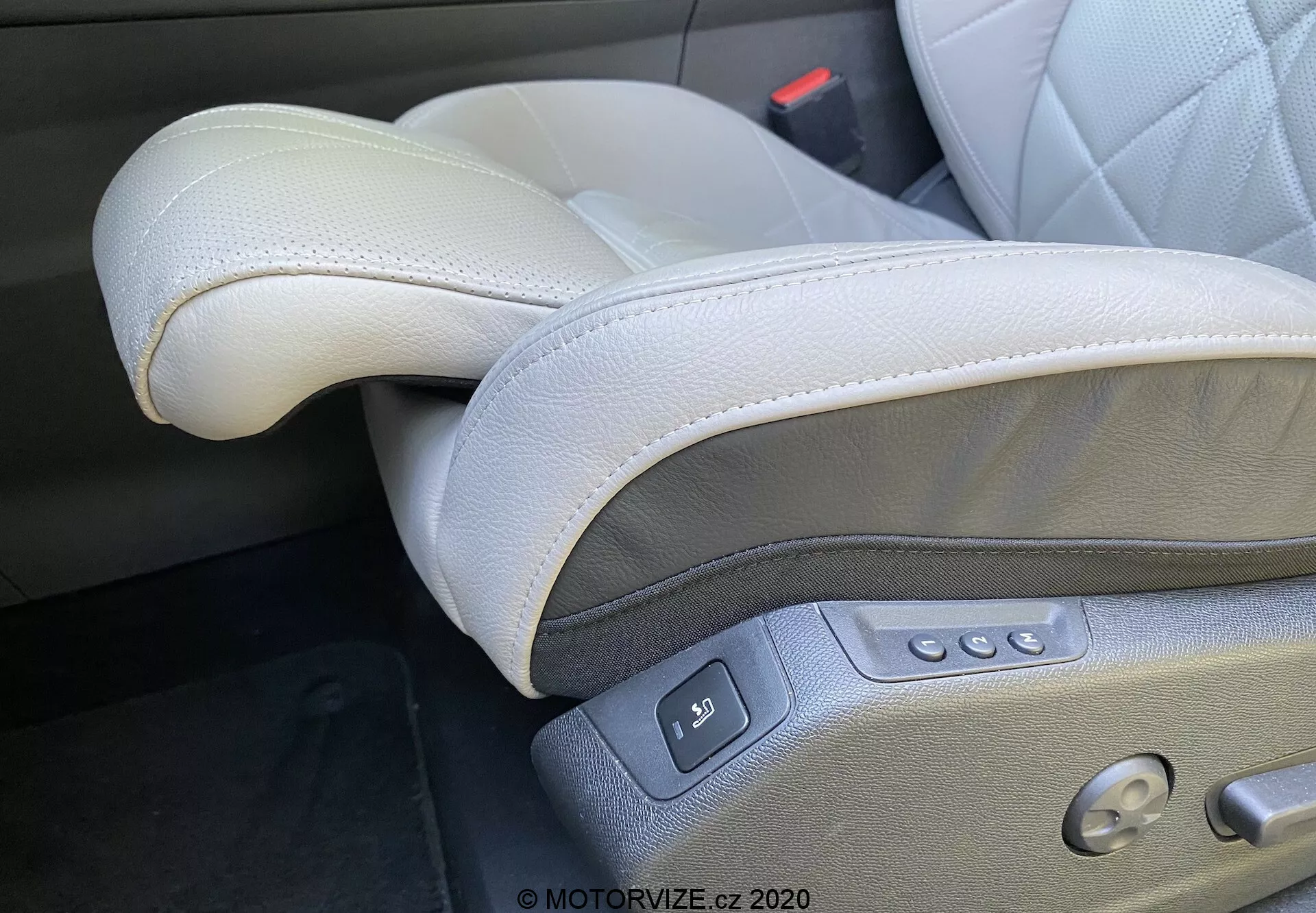 Bližnji posnetek notranjosti DS Automobiles DS-7 2018 (DS-7 Crossback), osredotočen na luksuzno svetlo sivo usnjeno voznikovo sedeže, ki se nahaja na levi strani v konfiguraciji z levo roko. Vidne funkcije vključujejo visokokakovostno usnje s šivi, nastavljiv vzglavnik, električne kontrole sedeža z več gumbi za drsenje, nagibanje, prilagajanje višine in ledveno oporo, pa tudi gumb za nastavitev pomnilnika in vidno zaponko varnostnega pasu, ki poudarjajo premium in ergonomski dizajn.
