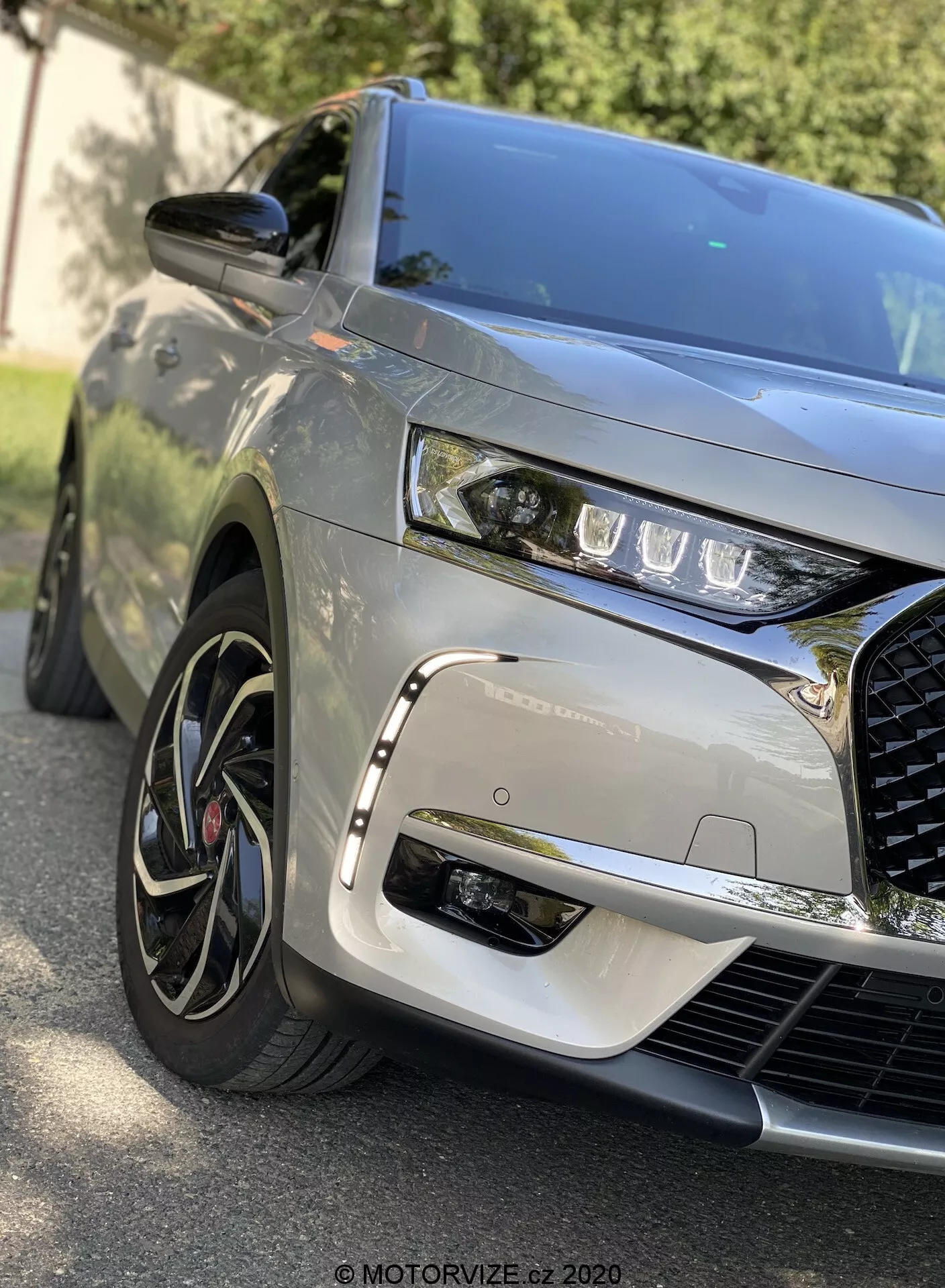 Od blizu pogled sprednjega kota DS Automobiles DS 7 Crossback 2018, zajemajoč levi sprednji del, prikazuje elegantne LED žaromete, izrazito in zapleteno rešetko, izklesano pokrov motorja, večkraka lita platišča z nizkoprofilnimi pnevmatikami, kovinsko barvo, ogledalo v barvi karoserije in dinamične odseve, ki nakazujejo zunanje okolje.