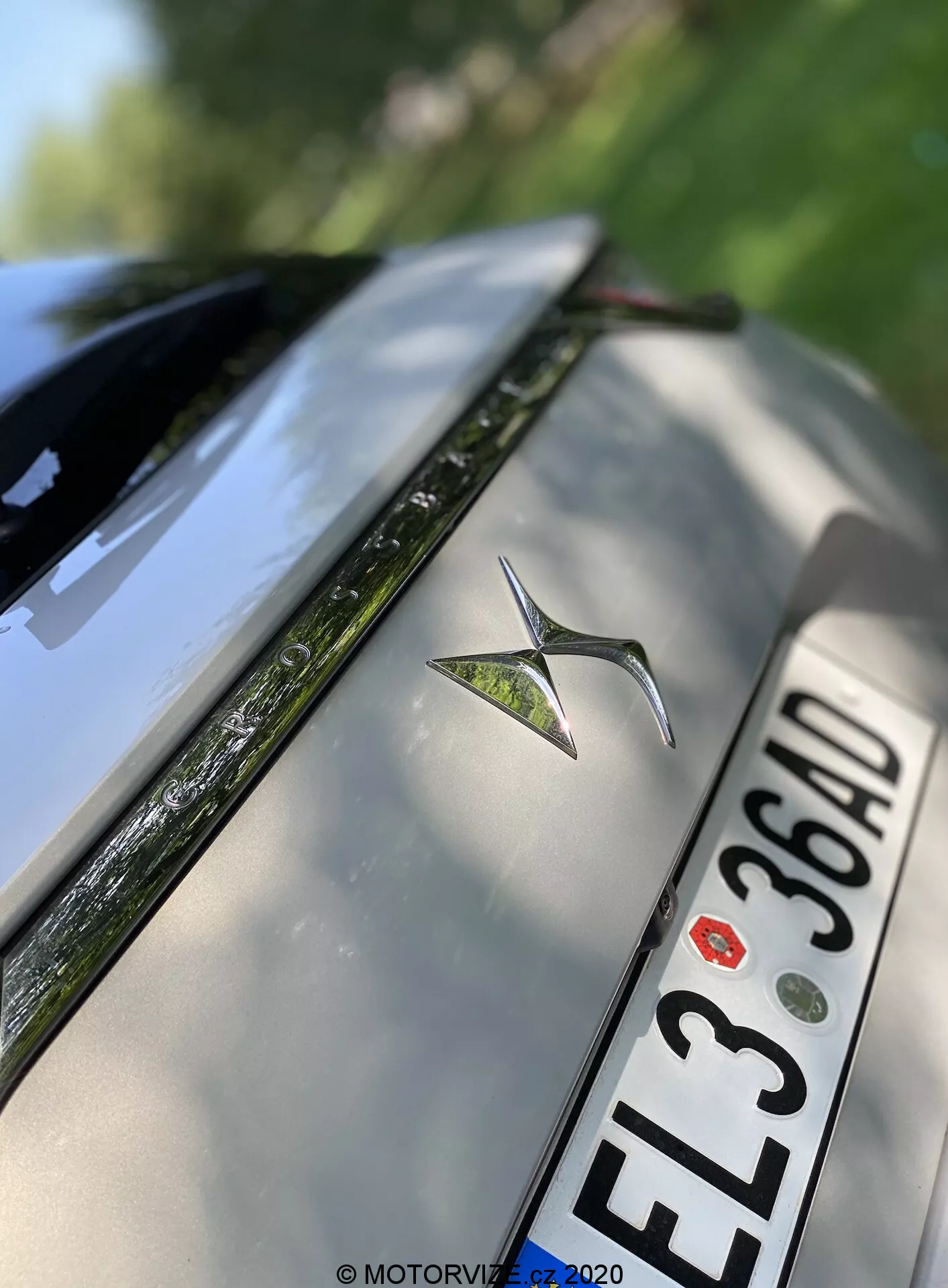 Zelo podroben poševni posnetek zadnjega dela srebrnega DS Automobiles DS 7 Crossback leta 2018. Slika poudarja kovinski emblem avtomobila, postavljen v sredino zadnjega pokrova, evropsko registrsko tablico s črnimi znaki 'EL3 36AD' pod emblemom in zgornji rob zadnjega odbijača. Del zadnjega vetrobranskega stekla z vidnim odmrzovalnikom ter bleščeče odsevne površine so tudi vidni. Plitvina globinske ostrine poudarja emblem, medtem ko nežno zamegli robove zadnjega pokrova in vetrobranskega stekla.