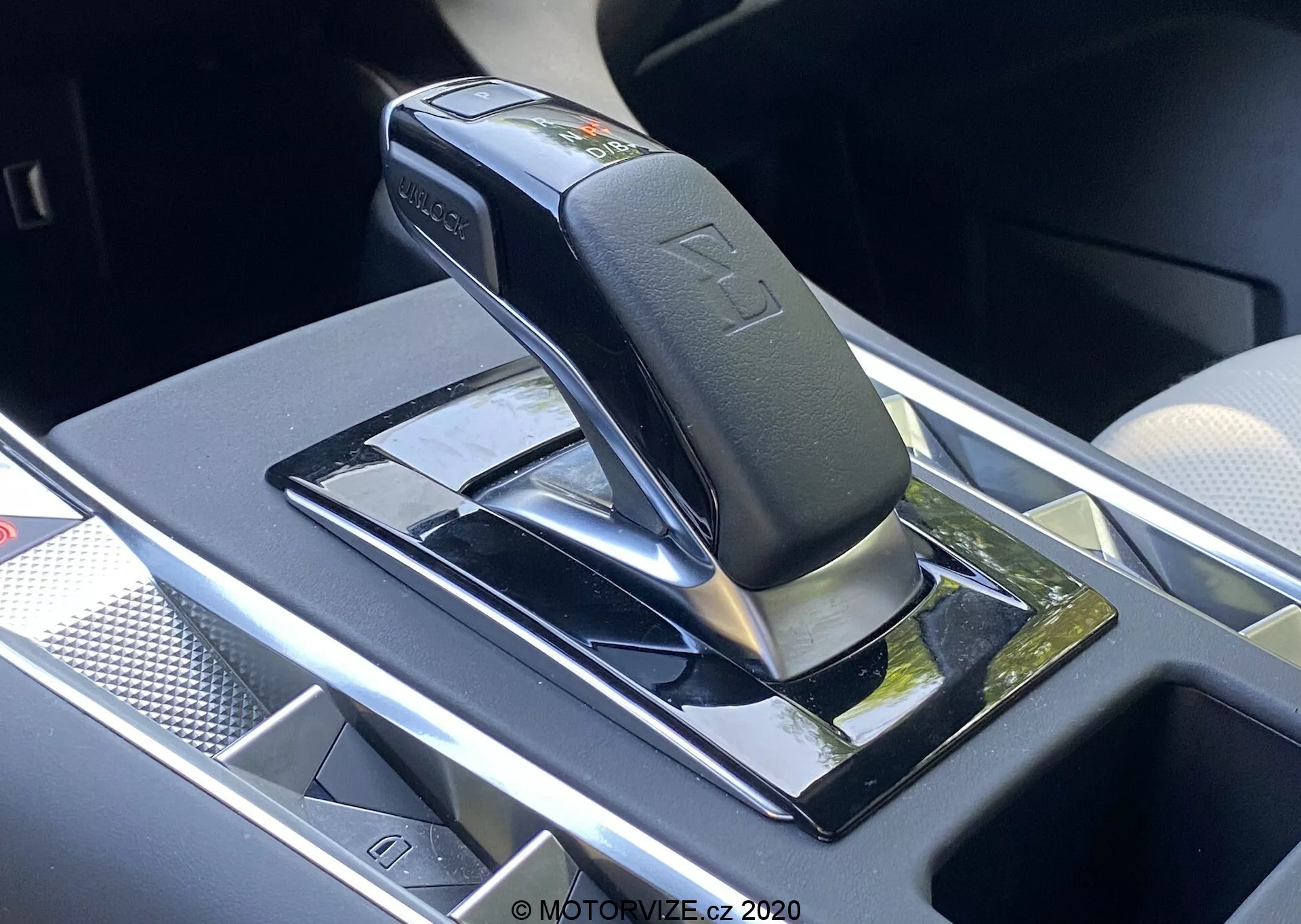 Pogled od blizu na avtomatsko prestavno ročico DS Automobiles DS-7 2018 (DS-7 Crossback), ki prikazuje sijočo črno ročico s srebrnimi poudarki, verjetno v parkirnem ali nevtralnem položaju, nameščeno na črni sijajni plošči z odsevnimi površinami. Značilnosti vključujejo vtisnjene simbole prestavnih mest, varnostni gumb na ročici in bližnji shranjevalni prostor z morebitnim brezžičnim polnilcem. Okoliško območje ima kromirane ali srebrne obrobe, kar pomeni sodobno in razkošno notranjost vozila.