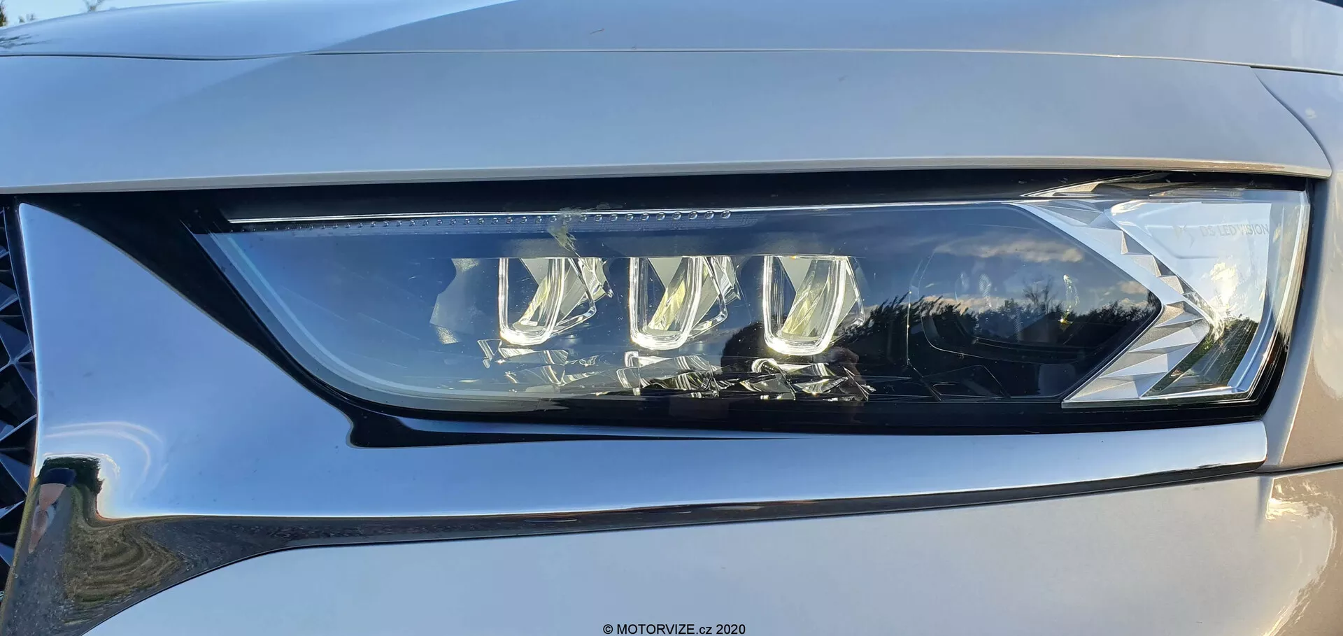 Odblizu levi žaromet DS Automobiles DS-7 Crossback iz rahlega kota, ki prikazuje sodobno LED zasnovo z več projektorskimi lečami in značilno dnevno LED lučjo, vgrajeno v prozorno, visoko kakovostno plastiko. Žaromet ponazarja napredno tehnologijo osvetlitve vozil, ki je sposobna zagotavljati funkcije kratkih in dolgih žarometov, s potencialnimi prilagodljivimi funkcijami za izboljšano vidljivost. Viden je tudi rahel odsev na sijajni površini in odtenek svetlo do srednje sive barve avtomobila.