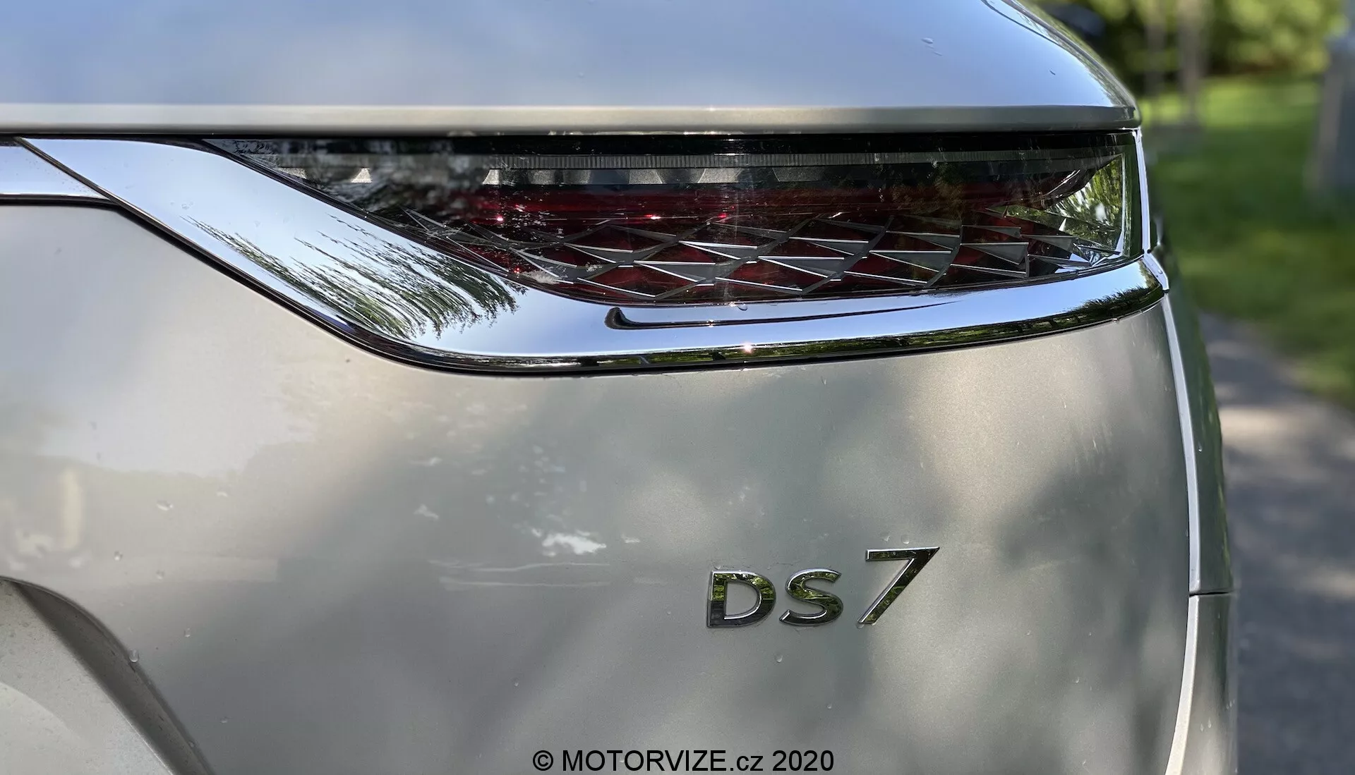 Pogled od blizu na zadnji del avtomobila DS Automobiles DS 7 Crossback 2018, posnet iz nizkega kota. Osredotočenost je na zadnja vrata z vidnimi, krepko izpisanimi črkami 'DS' nad oznako modela '7'. Nad imenom modela je vodoravno raztegnjena, ukrivljena zadnja luč z značilnimi LED elementi. Barva karoserije je kovinsko srebrna, oblikovan dizajn pa namiguje na sodoben in luksuzen videz. Fotografija poudarja blagovno znamko in dizajn zadnje luči ter subtilen odsev svetlega zunanjega okolja na površini avtomobila.