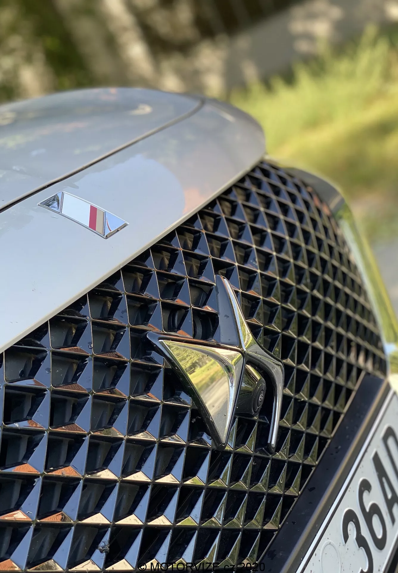 Pogled od blizu na prednji levi del DS Automobiles DS 7 Crossback 2018. Slika se osredotoča na luksuzno, zapleteno diamantno vzorčasto sprednjo rešetko z odsevnimi površinami, v središču s poudarjenim stiliziranim logotipom. Nad rešetko je na pokrovu motorja manjši emblem, ki označuje model višjega razreda. Ozadje je zamegljeno, kar poudarja dizajn rešetke in elemente blagovne znamke.