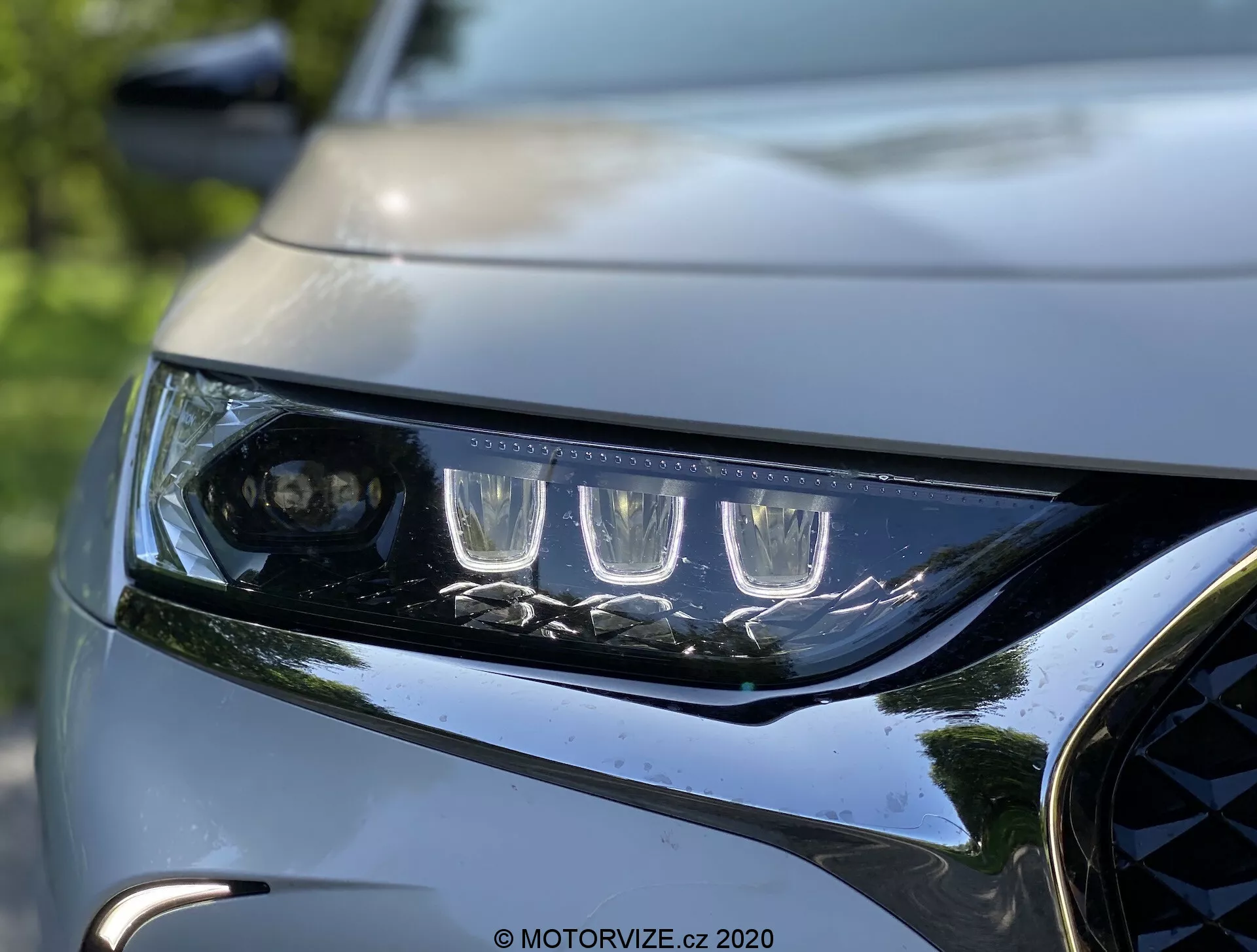 Zbližan posnetek levega žarometa in sprednje maske DS Automobiles DS 7 Crossback 2018. Žaromet ima značilne LED dnevne luči in lečo projektorja za glavni snop, obkrožen z več LED elementi. Elegantno in kotno ohišje žarometa prikazuje moderno aerodinamiko. Del temne, sijoče maske z kovinskimi poudarki in del oblikovane pokrove poudarjata dinamično, premijsko zasnovo vozila. Ozadje je zamegljeno, kar poudarja ostrino in dovršene podrobnosti osvetlitve in sprednje fasade avtomobila.