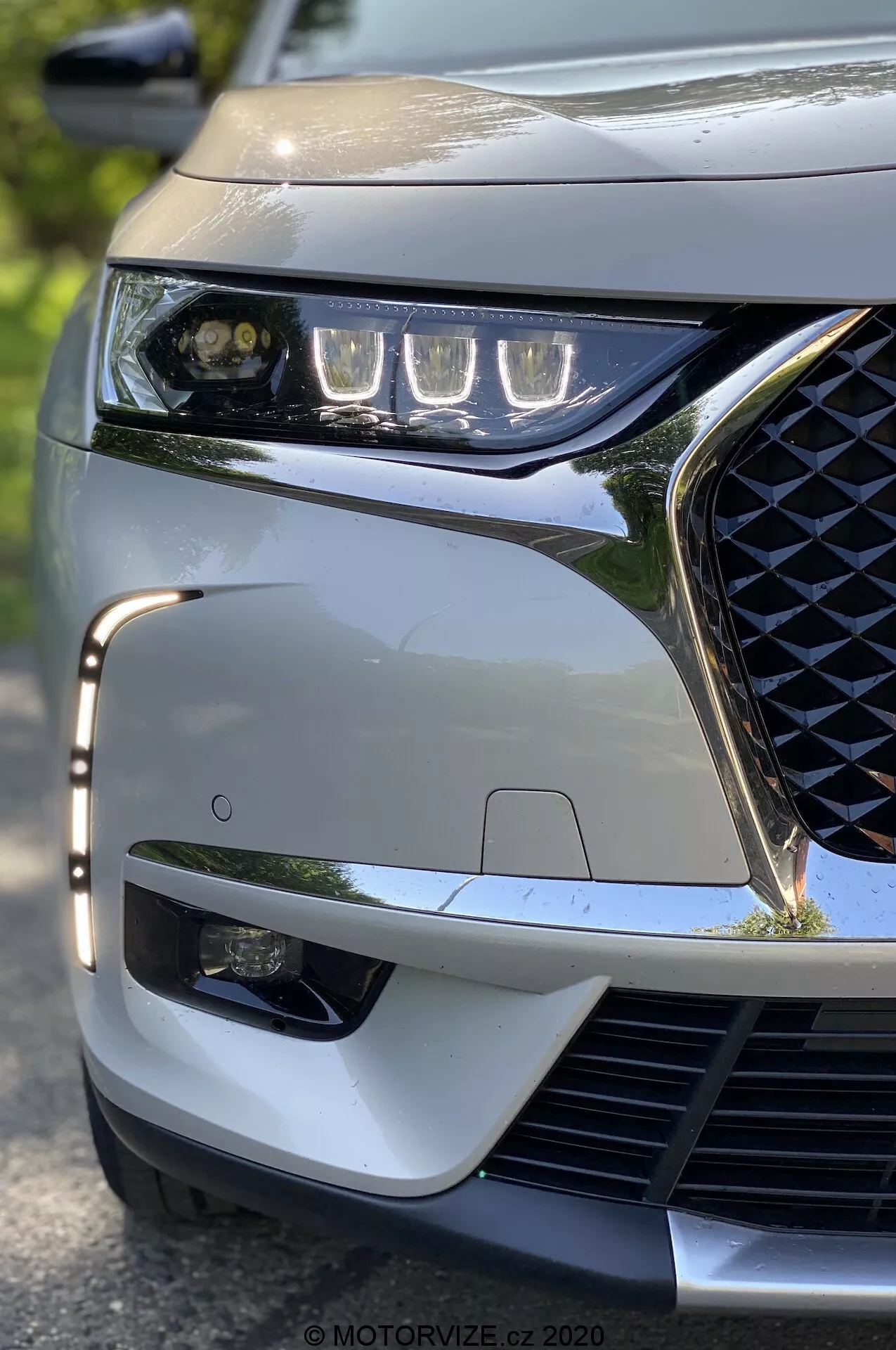 Približan posnetek iz nizkega kota na sprednji levi kot modela DS Automobiles DS 7 Crossback iz leta 2018. Poudarjene so elegantne LED luči, geometrijsko vzorec mreže, športni prednji odbijač in viden senzor ADAS, kar poudarja moderni tehnološki in estetski značaj vozila.
