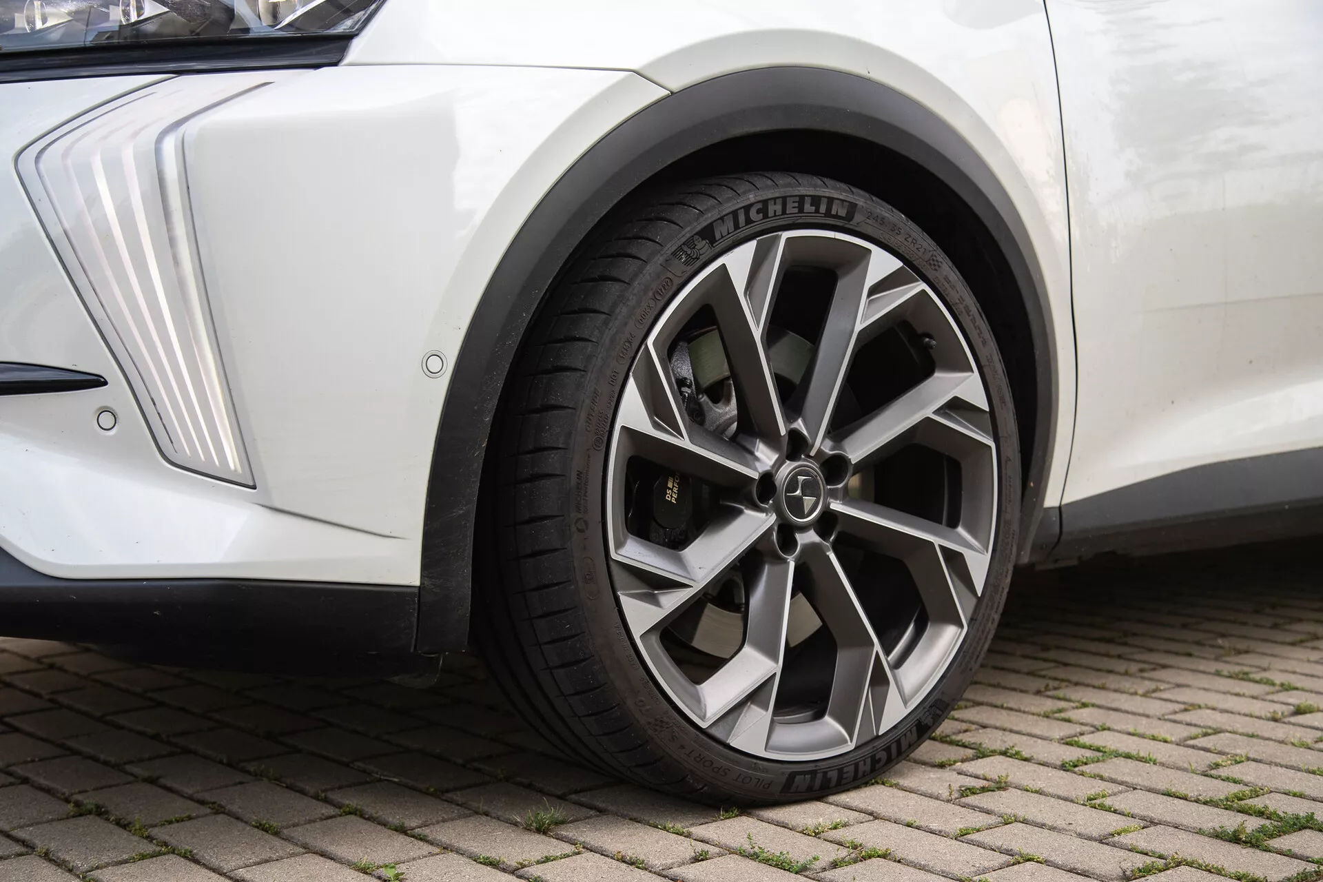 Pogled od blizu na sestavo sprednjega kolesa DS-Automobiles DS-7 2022 na površini iz prepletenih opek, z značilnostmi kovinskega zaključka večkrake litačne platišča z logotipom na sredinskem pokrovu, radialne pnevmatike Michelin z izrazitimi vzorci tekalne plasti, belo blatobrana z ostrim oblikovalskim črtami in črnim blatnikom, pod naravno svetlobo