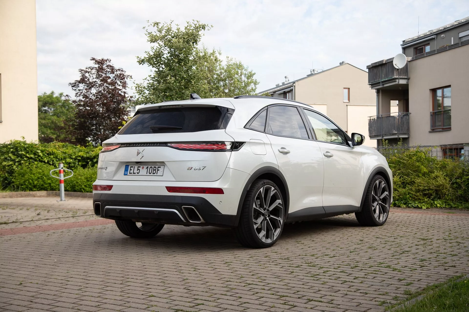 Zadnji tričetrtinski pogled na DS Automobiles DS 7 2022 v beli barvi, prikazuje stranski profil in zadnji del z značilnimi ozkimi horizontalnimi zadnjimi lučmi, značkami, visoko nameščenim spojlerjem, anteno v obliki morskega psa, senzorje za parkiranje, dvojnim izpušnim sistemom in velikimi liti kolesi z več kraki na stanovanjski ulici.