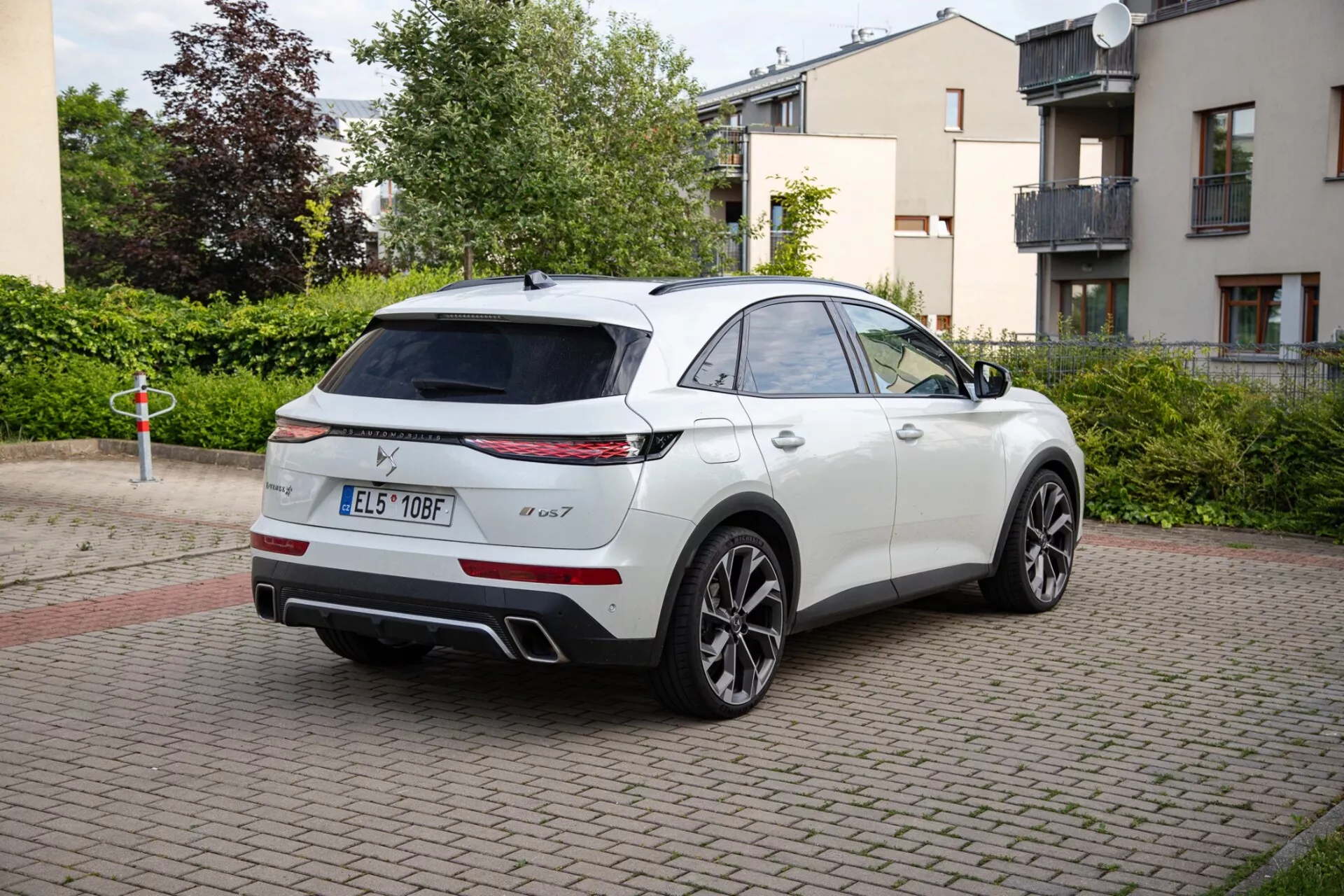 Beli SUV DS Automobiles DS-7 letnik 2022, parkiran na območju z opečnato površino, prikazuje LED zadnje luči, dvojni izpušni sistem in športna lita kolesa. Slika prikazuje vozilo s šik SUV dizajnom podobnim kupeju iz perspektive treh četrtin zadaj, poudarjajoč možno panoramsko streho in tehnološko dovršena ogledala.