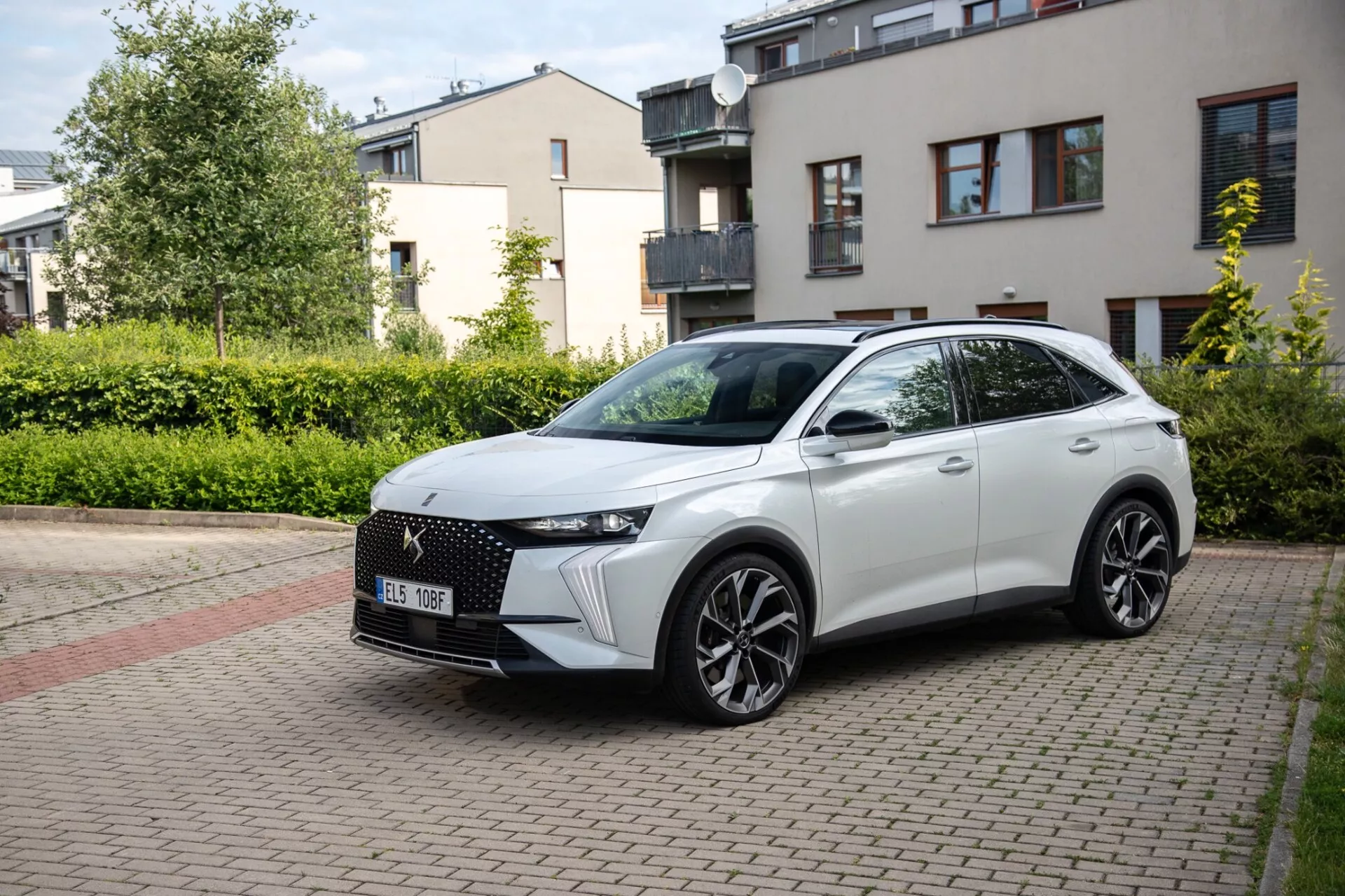 Tričetrtinski sprednji pogled na crossover DS Automobiles DS-7 2022, parkiran na opečnati stanovanjski ulici, ki prikazuje njegov sodoben dizajn, vključno z značilno sprednjo rešetko, elegantnimi LED žarometi, oblikovano pokrovom motorja, velikimi litoželeznimi kolesi in športnim stranskim profilom z integriranimi smerokazi na vratih montiranih stranskih ogledalih pod naravno dnevno svetlobo.