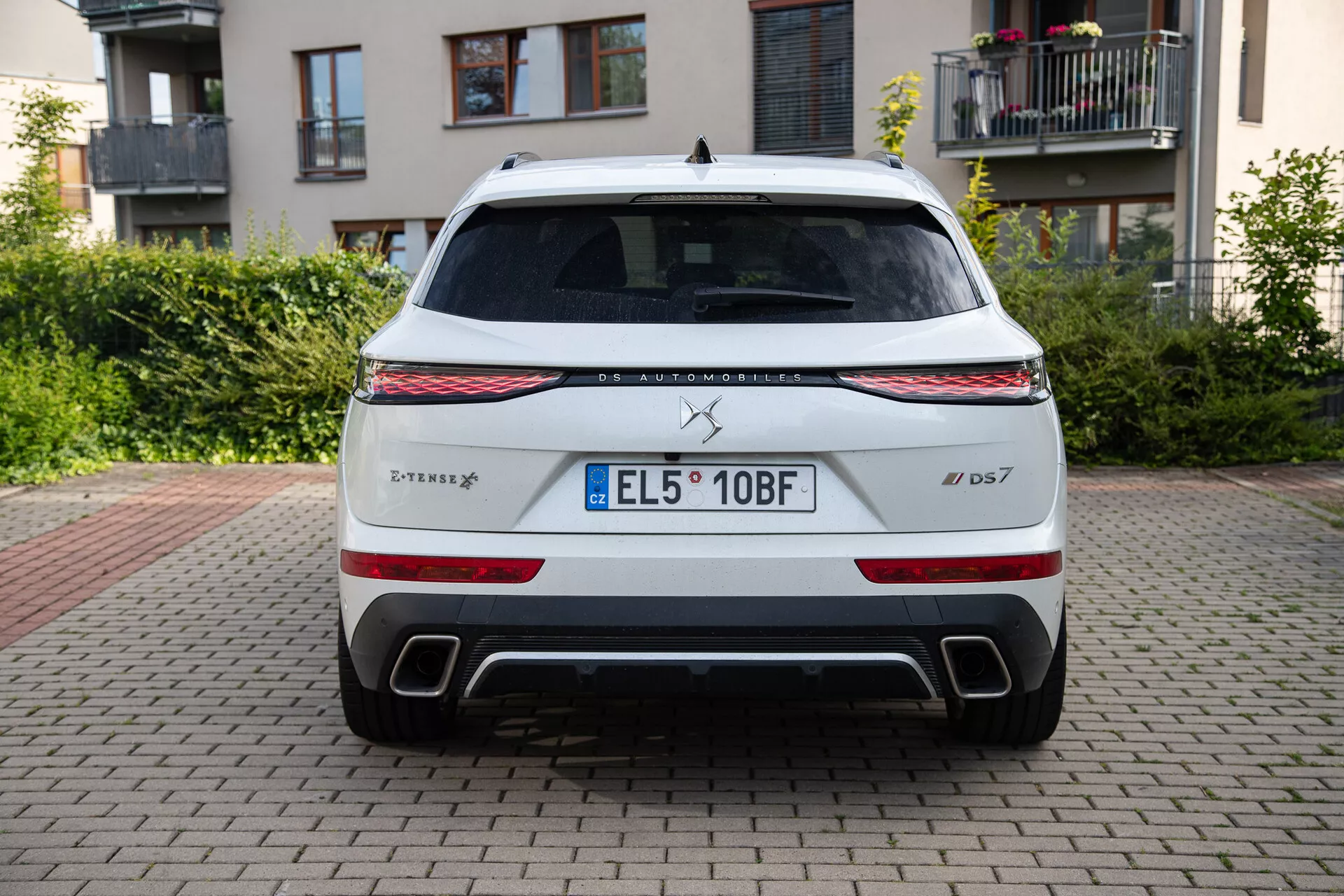 Zadnji pogled na DS Automobiles DS 7 2022, parkiran na opečnatem površju z zgradbami in zelenjem v ozadju, prikazuje LED svetlobni trak, zadnje luči, dvojne izpušne odprtine in registrsko tablico EU