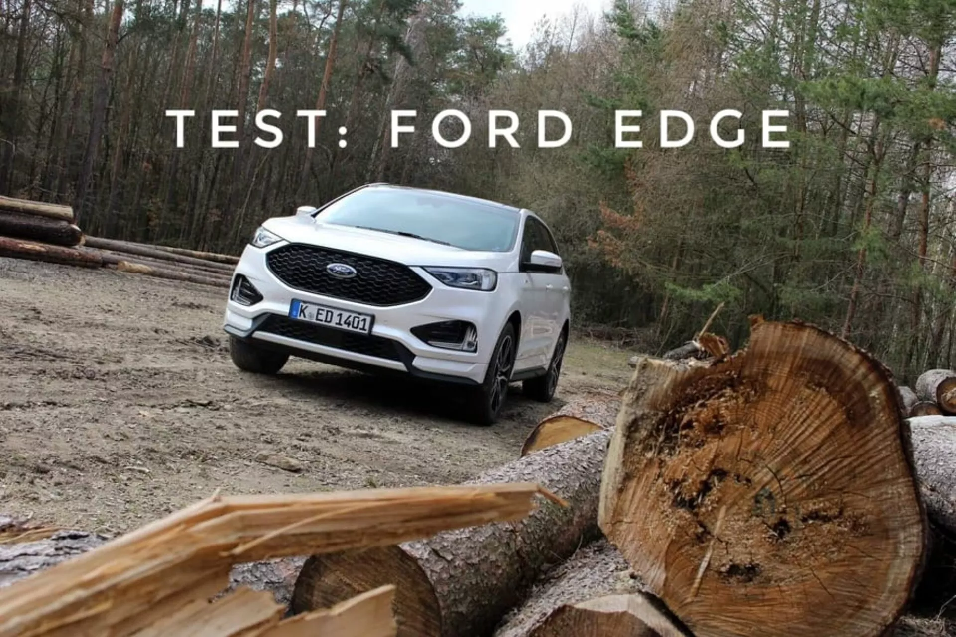 Beli Ford Edge 2018 prikazuje svoje zmožnosti za off-road z vidika tri četrt spredaj in stranske strani, poudarja LED dnevne luči, večkraka lita kolesa in dvobarvno barvanje, v naravnem ozadju z rezanimi hlodov lesa v ospredju