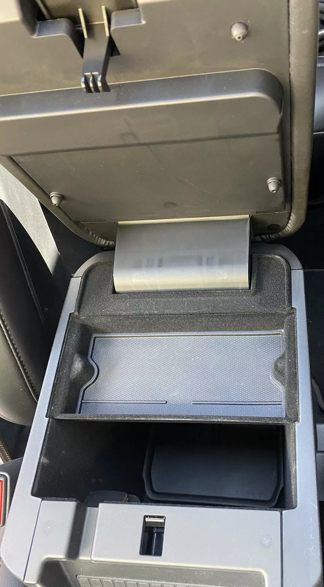 Od blizu notranjosti Ford Explorer 2019, detajl sredinske konzole, ki prikazuje odprt sredinski naslon za roke s premaknjeno sekundarno drsno pladnjo nazaj, razkrivajoč priključek USB in globlji prostor za shranjevanje v črni plastični obdelavi.