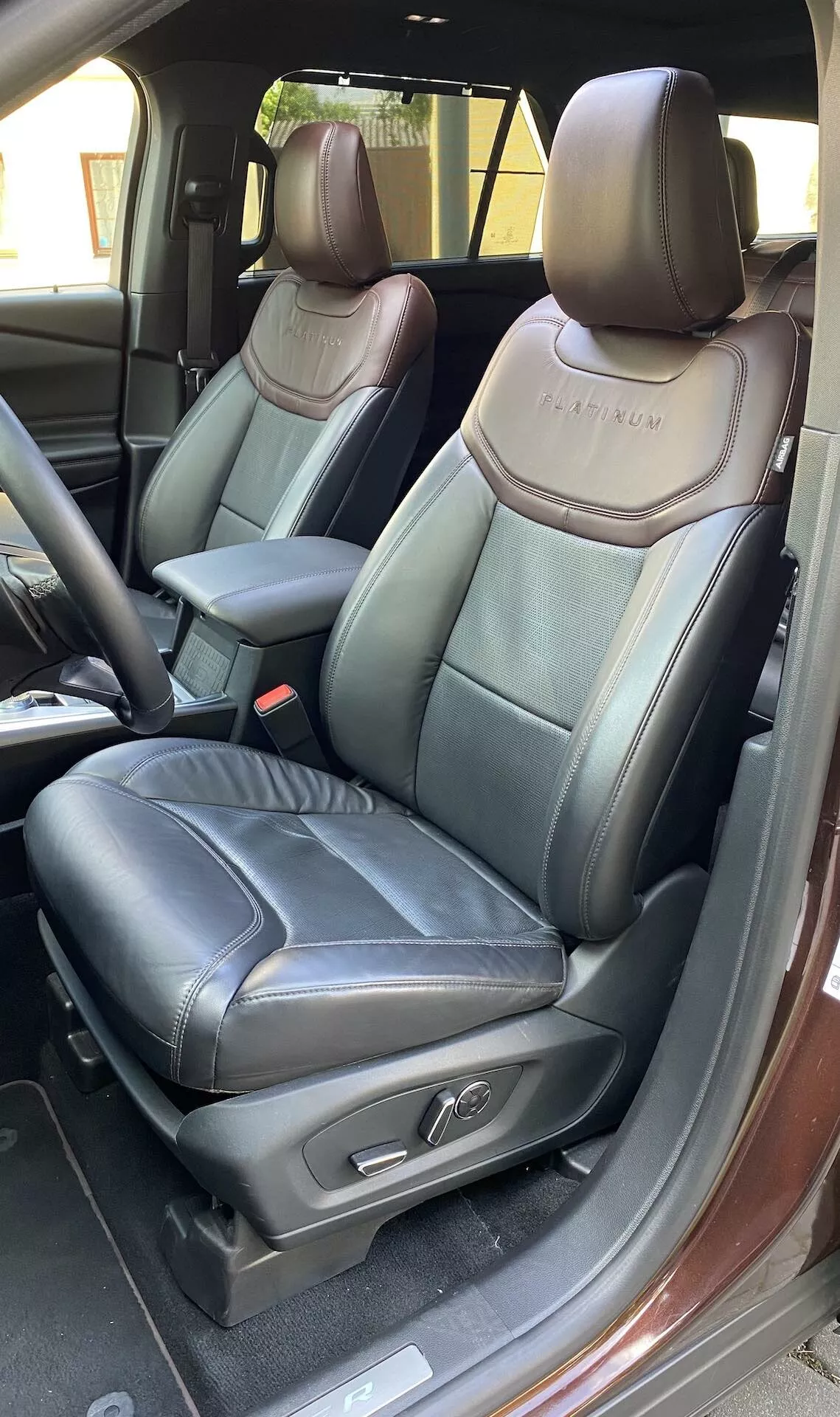 Podroben pogled na notranjost Ford Explorer 2019, osredotočen na voznikov sedež z odprtimi vrati. Fotografija poudarja črno usnjeno oblazinjenje s kontrastnimi šivi, vtisnjen logotip na naslonjalu sedeža, nastavljivo vzglavje, gumbe za nastavitev sedeža, naslon za roke na osrednji konzoli, vidni gumb za sprostitev varnostnega pasu, notranji mehanizem za zaklepanje vrat in kovinsko obrobo praga vrat.