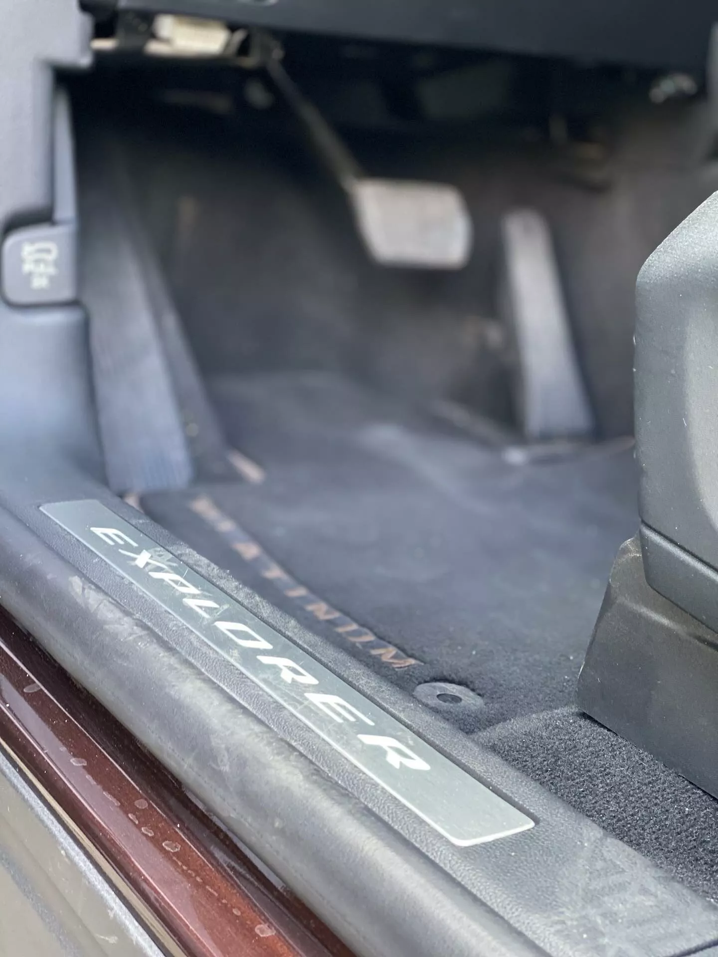 Bližnji posnetek notranjosti SUV Ford Explorer 2019, ki posebej prikazuje območje praga vrat z brušeno kovinsko ploščo z vgraviranim logotipom 'Explorer'. Slika poudarja prag, kjer vstopajo potniki, s temno preprogo, tesnili in mehko naravno osvetlitvijo.