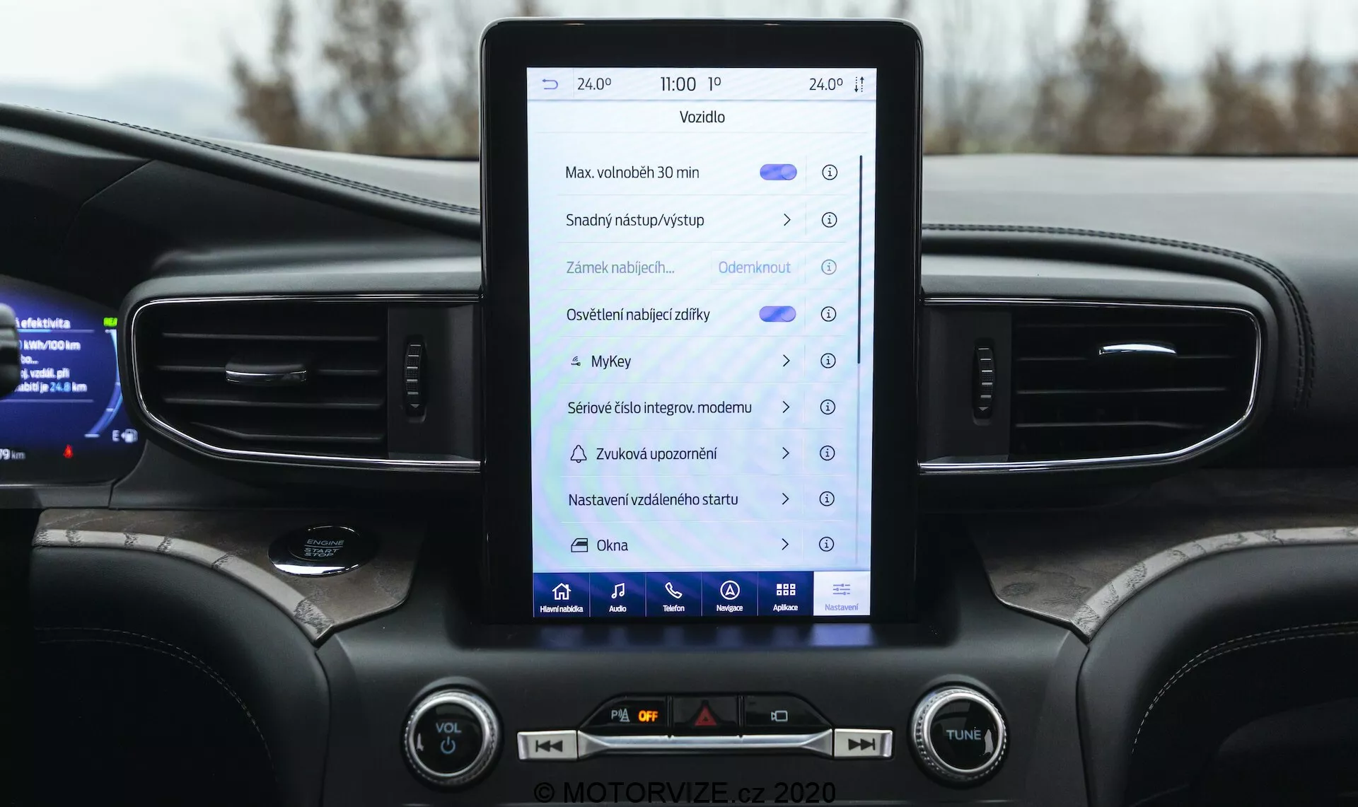 Povečan posnetek notranjosti Ford Explorer 2019, osredotočen na sredinsko konzolo in informacijsko-zabavni sistem. Navpično usmerjen zaslon na dotik prikazuje ikone in besedila, kar kaže na funkcije, kot so navigacija, nastavitve vozila, avdio-vizualni nadzor, Bluetooth in morda Apple CarPlay ali Android Auto. Armaturna plošča vključuje navpične zračne odprtine, ambientalno osvetlitev, fizične gumbe in gumbe za nadzor klime in glasnosti zvoka. Vidni so tudi zgornji robovi večfunkcijskega volana in delni digitalni instrumentni zaslon, ki prikazuje informacije o vozilu. Dizajn poudarja moderno, srednje ali premium vozilo s temno barvno shemo.