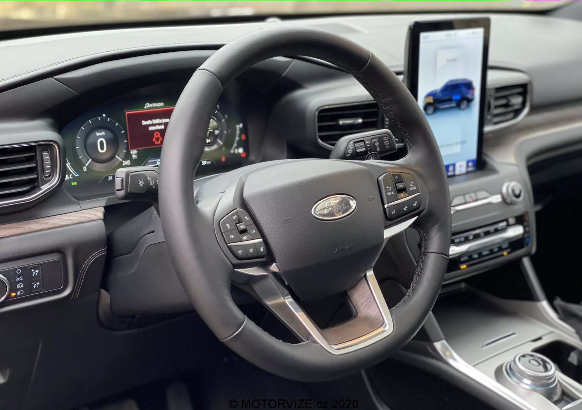 Pogled na notranjost Ford Explorerja 2019 iz perspektive sedeža voznika, z modernim triramenskim volanom s kromiranim poudarkom in večfunkcijskimi gumbi za tempomat, upravljanje medijev in glasovne ukaze. Za volanom je digitalni instrumentni grozd, ki prikazuje informacije o vozilu. Armaturna plošča vključuje velik zaslon na dotik, nagnjen proti vozniku za navigacijo in dostop do medijev, dopolnjen z rotacijskim izbirnikom prestav in visokokakovostnimi materiali.