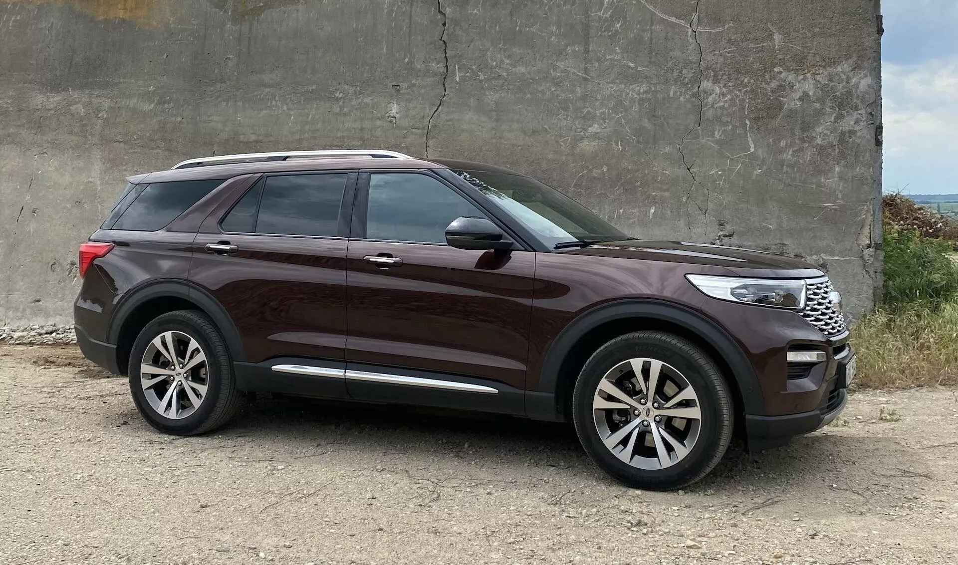 SUV Ford Explorer 2019 parkiran na grobem gramozu blizu betonske zgradbe, stranski profil poudarja oblikovne elemente, kot so velika lita platišča, visoka bočna linija, zatemnjena okna, kljuke v barvi karoserije, kromiran pas na oknih, elegantne karoserijske linije, integrirani smerniki v ogledalih, globoka kovinska barva in opazna oddaljenost od tal.
