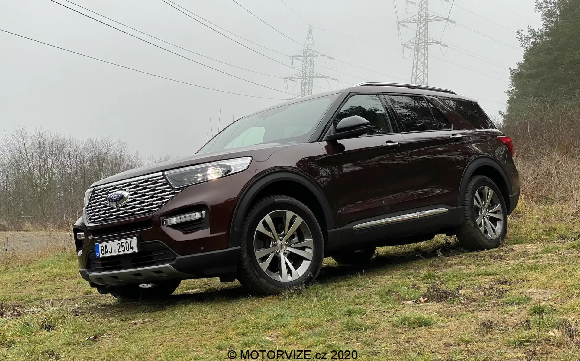 Slika temno obarvanega SUV-ja Ford Explorer 2019 parkiranega na neenakomernem travnatem terenu, ki poudarja njegovo robustnost in terenske zmogljivosti. Posneto iz sprednjega tričetrtinskega kota, SUV jasno prikazuje oblikovalske elemente, kot so izrazita maska z geometričnim vzorcem, znak znamke, široki zračni dovod, meglenke, elegantni objemajoči LED žarometi s kovinskimi poudarki ter večkraka lita platišča. Funkcije vključujejo tudi zrcala v barvi karoserije z integriranimi smerniki in strešne nosilce, kar nakazuje na praktičnost in prefinjenost.