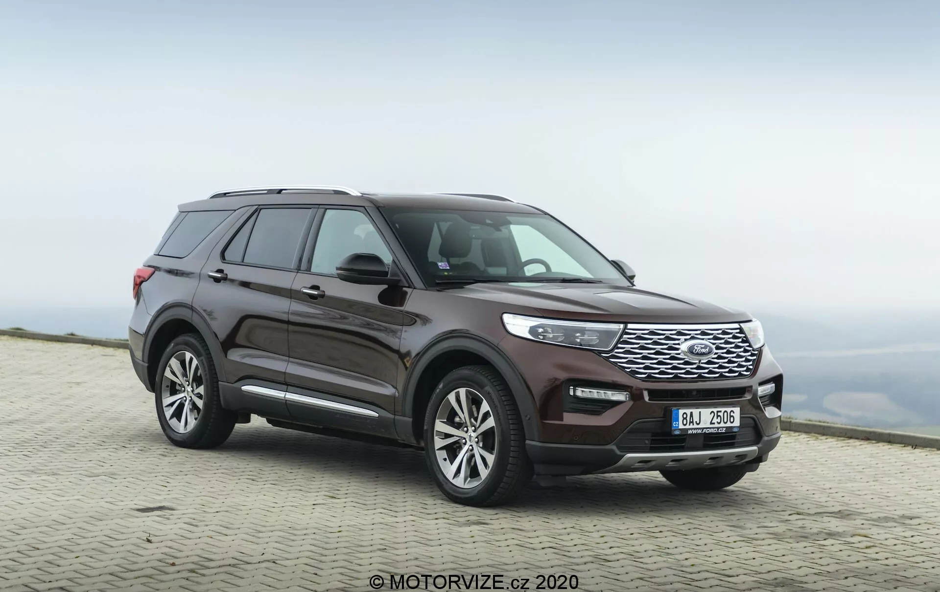 Slika SUV Ford Explorer 2019, zajeta iz trikotnega sprednjega kota, poudarja bočni profil in prednjo stran vozila. Nizka kamera poudarja robustno držo in izrazito sprednjo mrežo vozila. Vidna je celotna dolžina vozila, ki prikazuje oblikovne linije, obliko koles in obrise vrat in blatnikov. Kolesa so rahlo obrnjena v desno, kar daje občutek gibanja. Izpostavljene značilnosti vključujejo sprednjo mrežo z vodoravnimi letevmi, elegantne žaromete, ki se integrirajo z mrežo, in sprednji odbijač z vidnimi senzorji. Stranska ogledala verjetno vključujejo integrirane smerne kazalce, linija strehe pa se rahlo spušča proti zadnjemu delu z vidnimi strešnimi nosilci. Barva barve je globoko kovinsko bordo. Vozilo je parkirano na tlakovanem površju z zamegljenim naravnim ozadjem, kar kaže na visoko nadmorsko višino z oblačnim nebom.