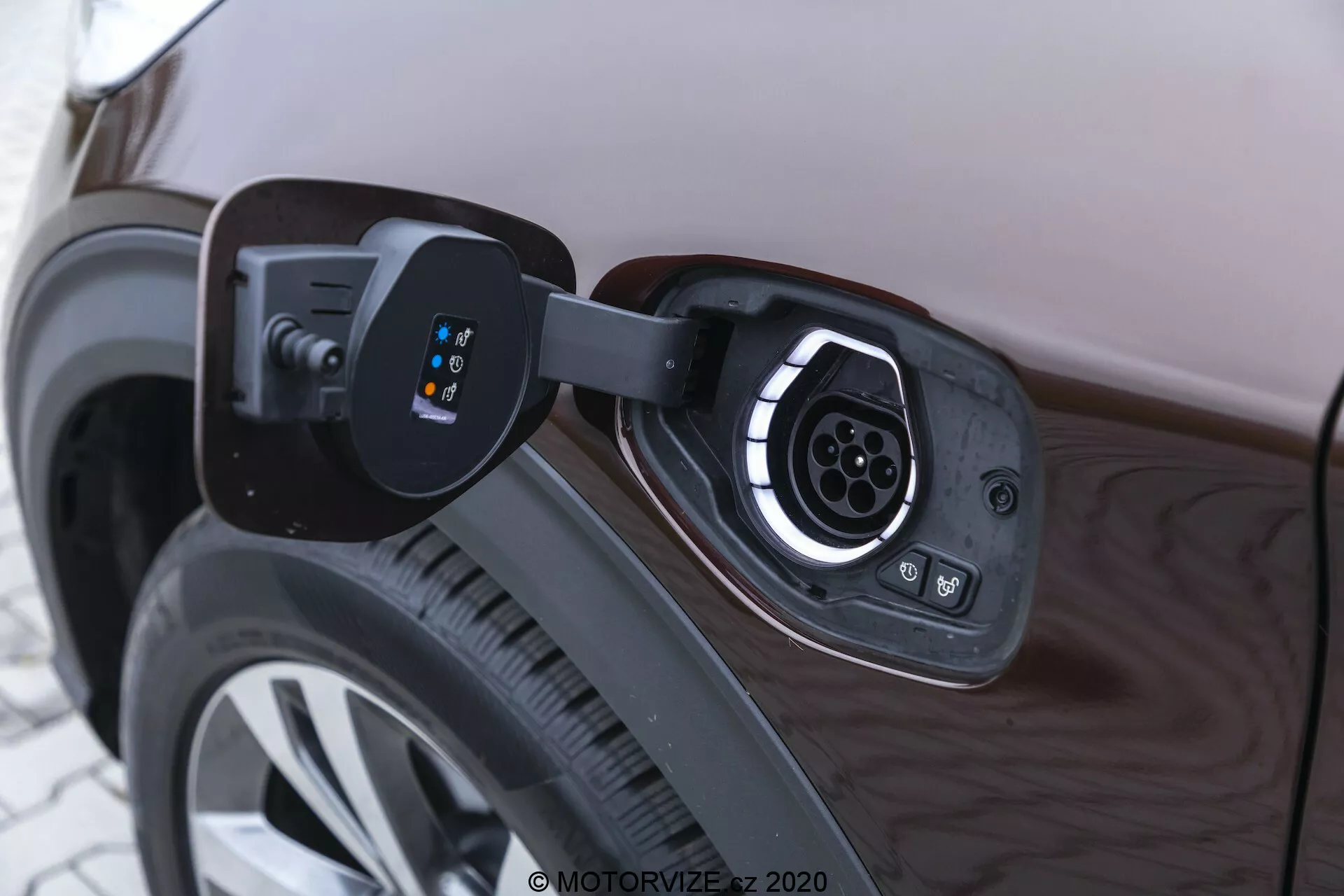 Povečan pogled na polnilni priključek električnega vozila na zadnjem četrtinskem panelu blizu zadnjega kolesa. Odprti pokrov razkriva vtičnico z več konektorji, značilnimi za hitro polnjenje tipa 2 ali podobnega standarda, s krožnim vzorcem zatičev. LED indikatorji na levi strani vtičnice prikazujejo stanje polnjenja. Slika se osredotoča na tehnologijo polnilnega sistema, stranski panel avtomobila in platišče kolesa pa odražata ambientalno svetlobo.