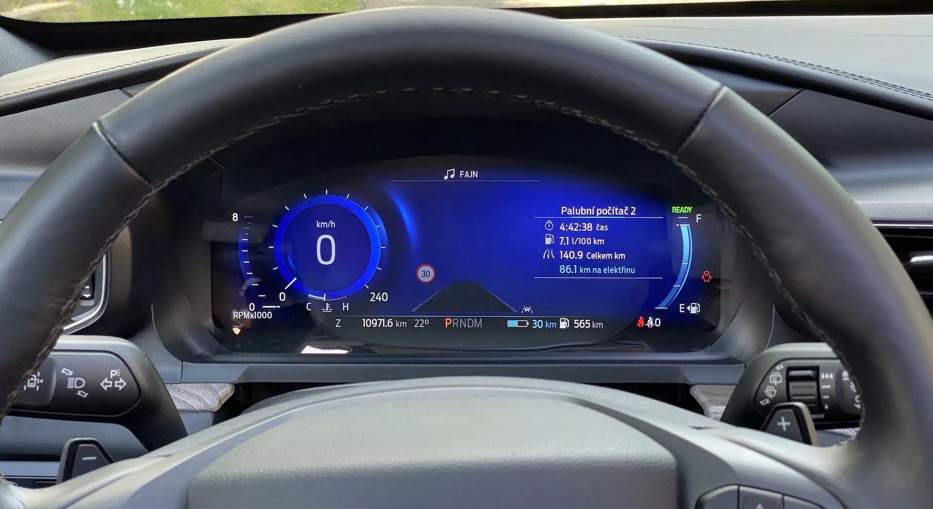 Notranji pogled na Ford Explorer 2019 iz voznikove perspektive, osredotočen na centralno postavljen volan in digitalno armaturno ploščo za njim. Volan ima različne kontrolne gumbe na obeh krakih. Za njim je popolnoma digitalni instrumentni zaslon, ki prikazuje informacije, kot so hitrost, merilnik moči, vozni doseg, raven goriva, zunanja temperatura in branje učinkovitosti. Armaturna plošča prikazuje tudi čas in datum, in je zasnovana z modernimi, tehnološko naprednimi elementi v modri in beli barvi. Volan je iz črnega usnja s šivi.