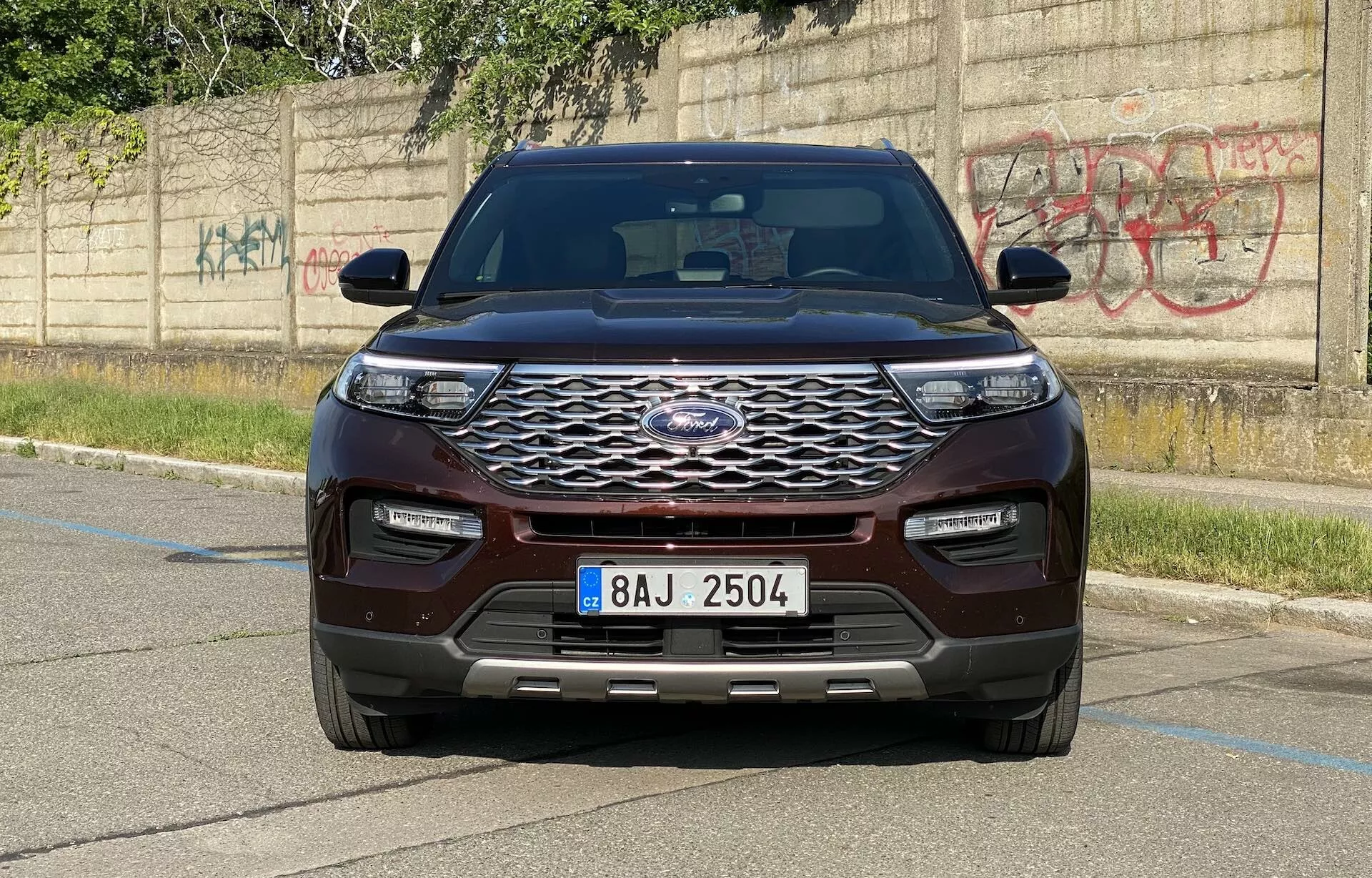 Sprednji pogled SUV-ja Ford Explorer 2019, zajet iz nivojskega kota, ki poudarja sodoben dizajn vozila. Izrazit sprednji del vozila vključuje velik kromiran rešetko s šestkotnim vzorcem in centralno postavljenim emblemom proizvajalca. Elegantni, vodoravno podaljšani žarometi LED in odbijač v barvi karoserije s spodnjimi dovodi zraka in parkirnimi senzorji poudarjajo tehnološke napredke. Pokrov motorja prikazuje oblikovan dizajn z dvignjenimi črtami, medtem ko blatniki in stranska ogledala z vgrajenimi smerniki dodajajo mišičast videz. SUV, pobarvan v bogato kostanjevo ali temno rdečo barvo, je parkiran na tlakovani površini z betonskim robnikom in steno, pokrito z grafiti v ozadju, kar poudarja urbano okolje.