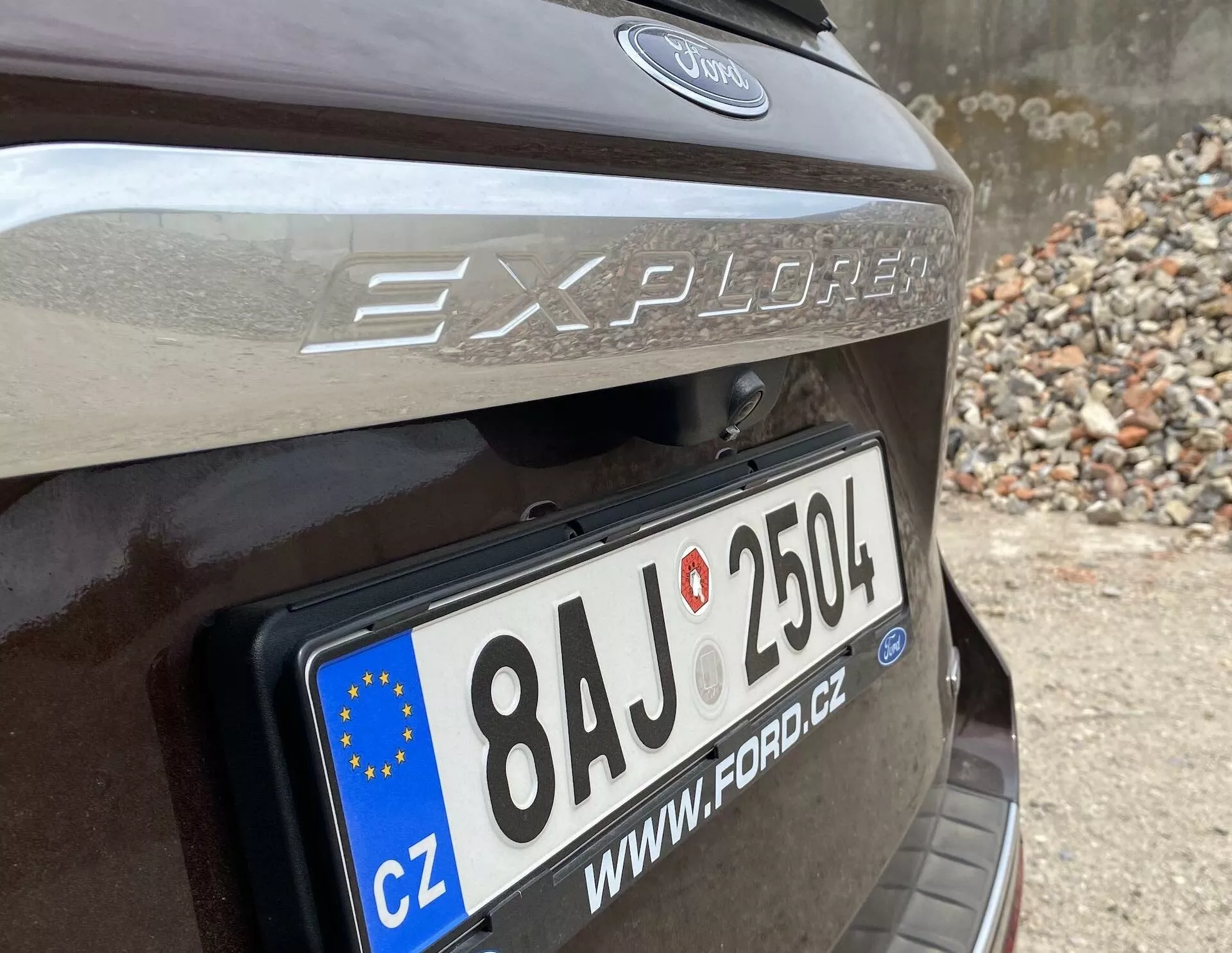 Bližnji pogled na zadnji del vozila Ford Explorer 2019, osredotočen na območje zadnjih vrat z jasno vidno registrsko tablico Evropske unije s številko '8AJ 2504' in oznako države 'CZ'. Nad registrsko tablico sta vidna oznaka modela 'Explorer' in logotip Forda, nad registrsko tablico pa je viden odsevni trak. Avto je temno rjave ali temno sive barve in parkiran na območju z kamenjem ali ruševinami.