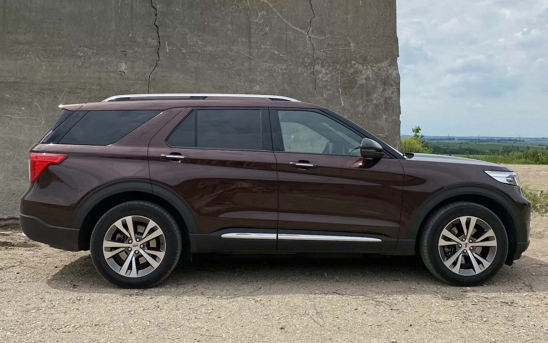 Bočni pogled na sodoben srednje velik SUV Ford Explorer 2019 z metalno rjavo barvo, večkrakimi litimi platišči, zatemnjenimi okni in strešnimi nosilci. Vozilo je parkirano na gramozni površini s preprosto steno in odprtim nebom v ozadju, ujeto v svetli opoldanski svetlobi.