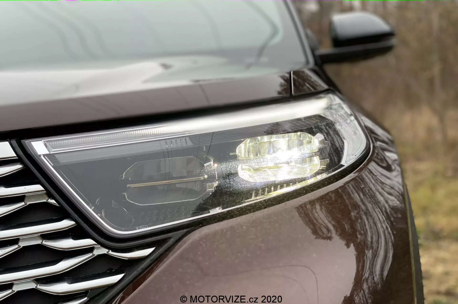 Drobni stranski pogled na sprednji del Ford Explorerja 2019. Izrazit sodoben žaromet, ki nakazuje LED ali HID tehnologijo z vključenimi dnevnimi lučmi. Delno vidna mreža z značko kovinskega zaključka, nameščena pod nizkim kotom. Avto ima temen, odseven lak, kar kaže na kovinski ali biserni zaključek, v zunanjih pogojih.