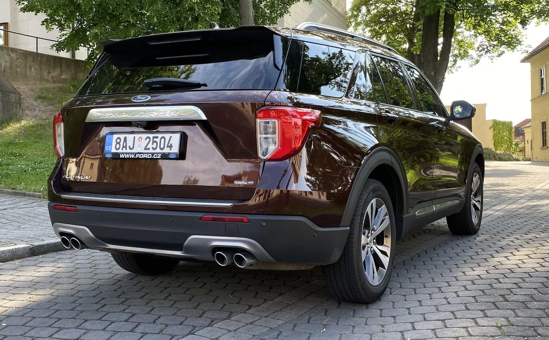 Sodobni SUV Ford Explorer 2019 z zadnje tričetrtinske perspektive, ki prikazuje zasnovo zadnjih luči, zadnjega odbijača s parkirnimi senzorji, dvojne izpušne cevi, aluminijasta platišča z več prečkami in temno kovinsko barvo, parkiran na tlakovanem površju v urbanem okolju z rastlinjem in zgradbami v ozadju.