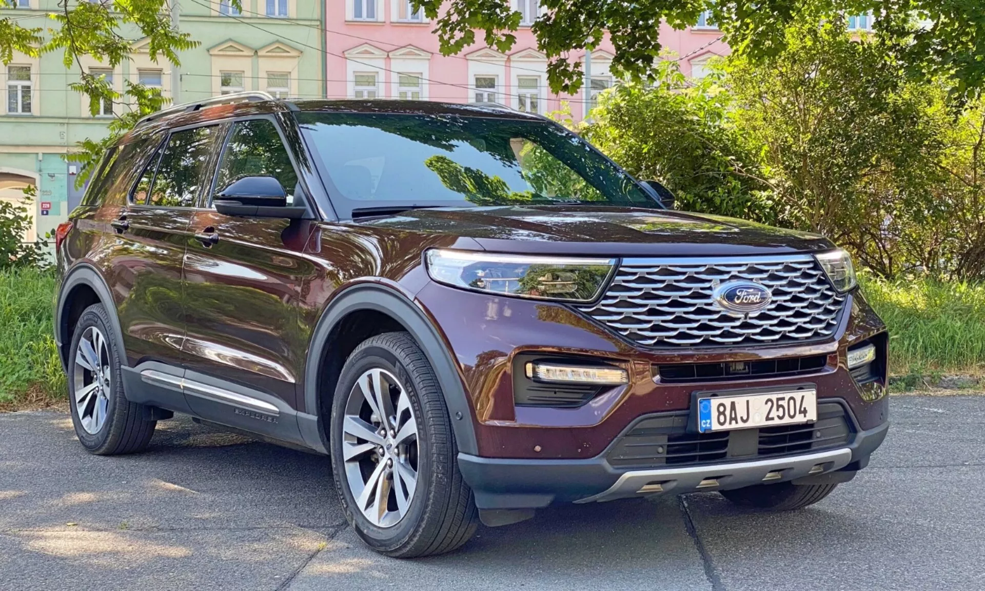 Tričetrtinski sprednji pogled na sodoben, rjav SUV Ford Explorer 2019, ki prikazuje njegov stranski profil in sprednjo fasado. Vozilo je parkirano ob mestni cesti z zgradbami in drevesi v ozadju. Vidne ključne lastnosti vključujejo izrazito sprednjo masko z vodoravnimi letvicami in osrednjim emblemom, moderne LED dnevne luči, meglenke, integrirane v spodnji odbijač, velika večkraka aluminijasta platišča, ogledala z vgrajenimi smerniki, zatemnjena okna, strešne nosilce, oblikovane linije karoserije in dvignjeno oddaljenost od tal.