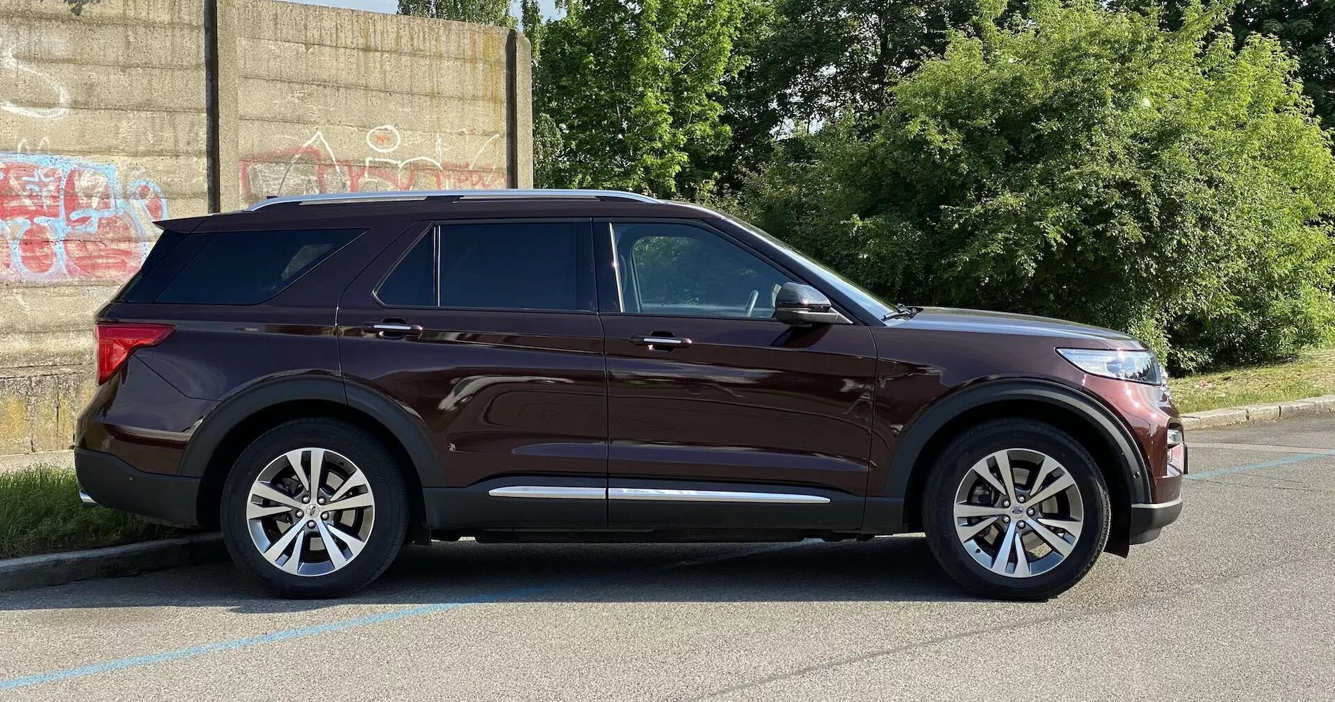 Sodobni SUV Ford Explorer 2019 iz stranskega pogleda, parkiran na tlakovanem območju poleg robnika z zidom, prekritim z grafiti v ozadju, kar nakazuje urbano okolje. Značilnosti vključujejo elegantne žaromete, izrazito značilno linijo, stranska ogledala s smerniki, petkraka alu platišča, kromirane detajle okoli oken, strešne nosilce, zatemnjena zadnja okna, ročaje vrat v barvi karoserije in zadostno oddaljenost od tal. Fotografija ob naravni dnevni svetlobi prikazuje celoten profil vozila.