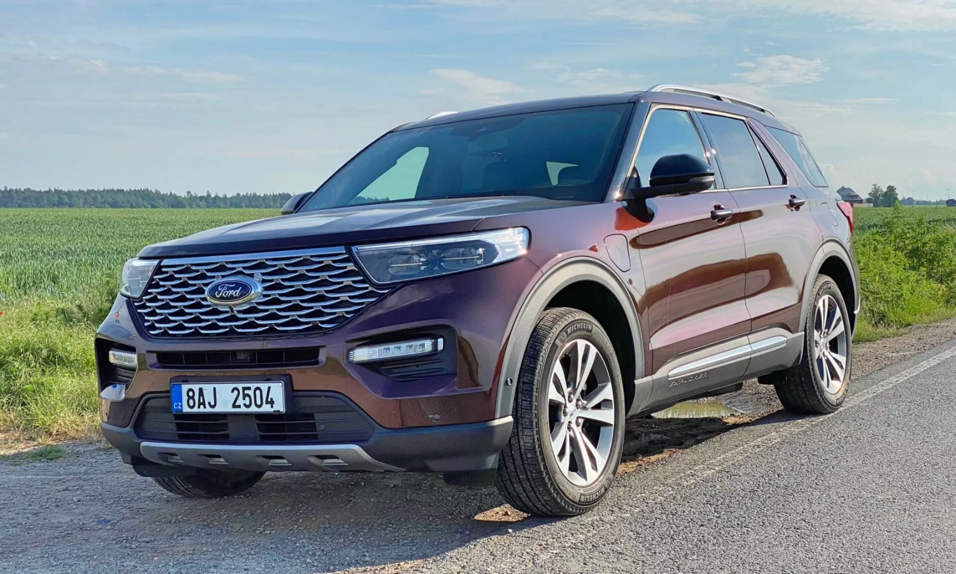 Videnje s treh četrtin spredaj na Ford Explorer SUV 2019. Slika prikazuje prednjo rešetko vozila z mrežastim vzorcem, elegantnimi podolgovatimi žarometi z videnimi dnevno-led svetilkami, oblikovan pokrov motorja z dvignjenimi obrisi, lita platišča z večkrakim dizajnom ter barvno usklajenimi tačnimi ogledali z vgrajenimi smernimi svetili. Okna imajo kromirano ali srebrno obrobo. Vozilo je kovinski kostanjev in je postavljeno na asfaltirano cesto v travnati, potencialno podeželski okolici.