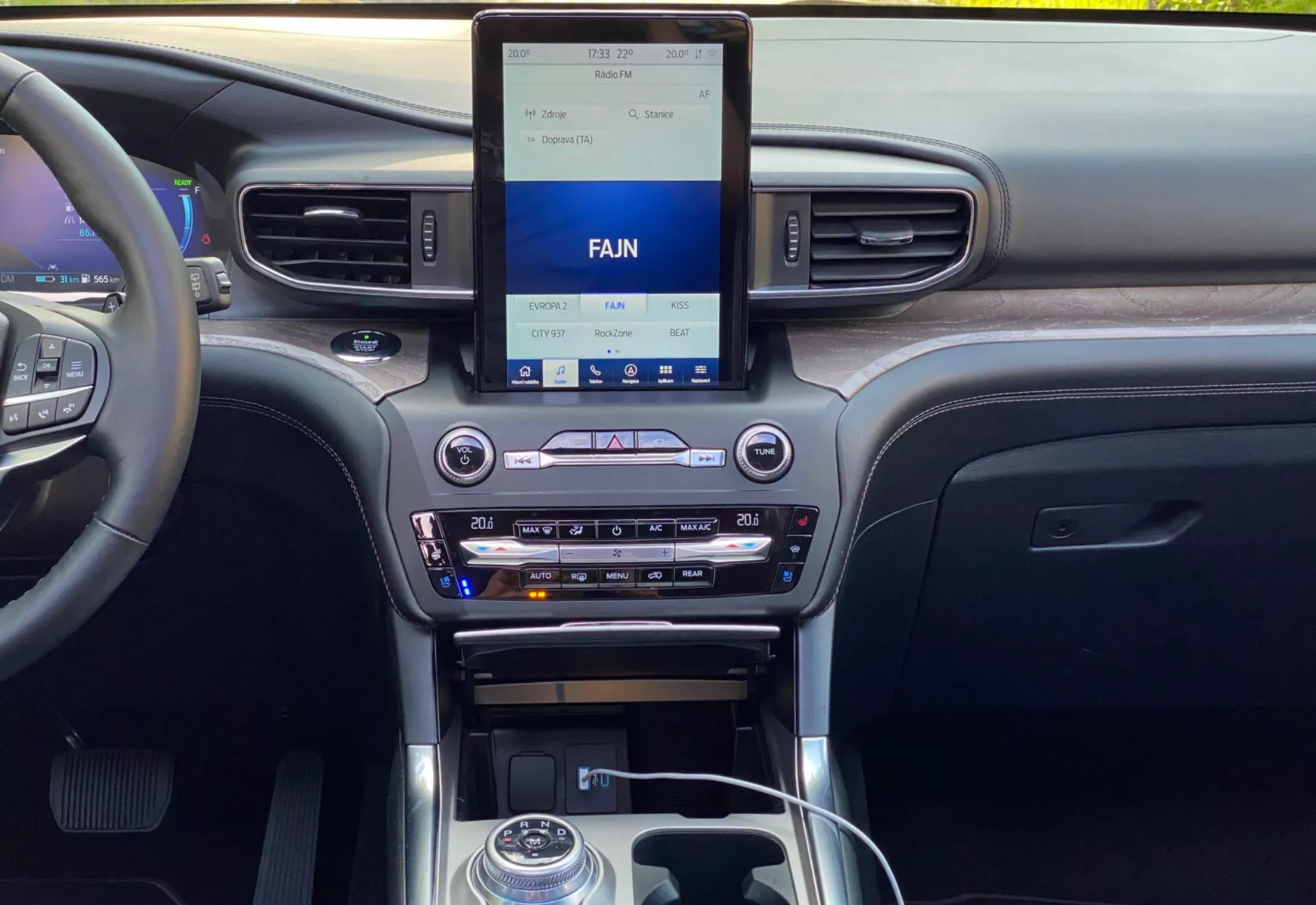Notranjost Ford Explorer 2019, ki prikazuje sodobno osrednjo armaturno ploščo iz perspektive prednjega ali zadnjega sedeža. Armaturna plošča vsebuje navpični infotainment zaslon na dotik s spodnjimi kontrolami klimatske naprave, vključno z nastavitvami dvojnega klimatskega sistema, režo za CD in možnostmi za vnos medijev. Osrednja konzola vključuje prestavno ročico s kovinskim poudarkom, kontrolne gumbe, vrtljiv gumb in držala za pijače. Materiali nakazujejo mešanico lesenega vzorca, kovinskih zaključkov in črne plastike, kar poudarja tehnologijo in kakovost notranje opreme vozila.