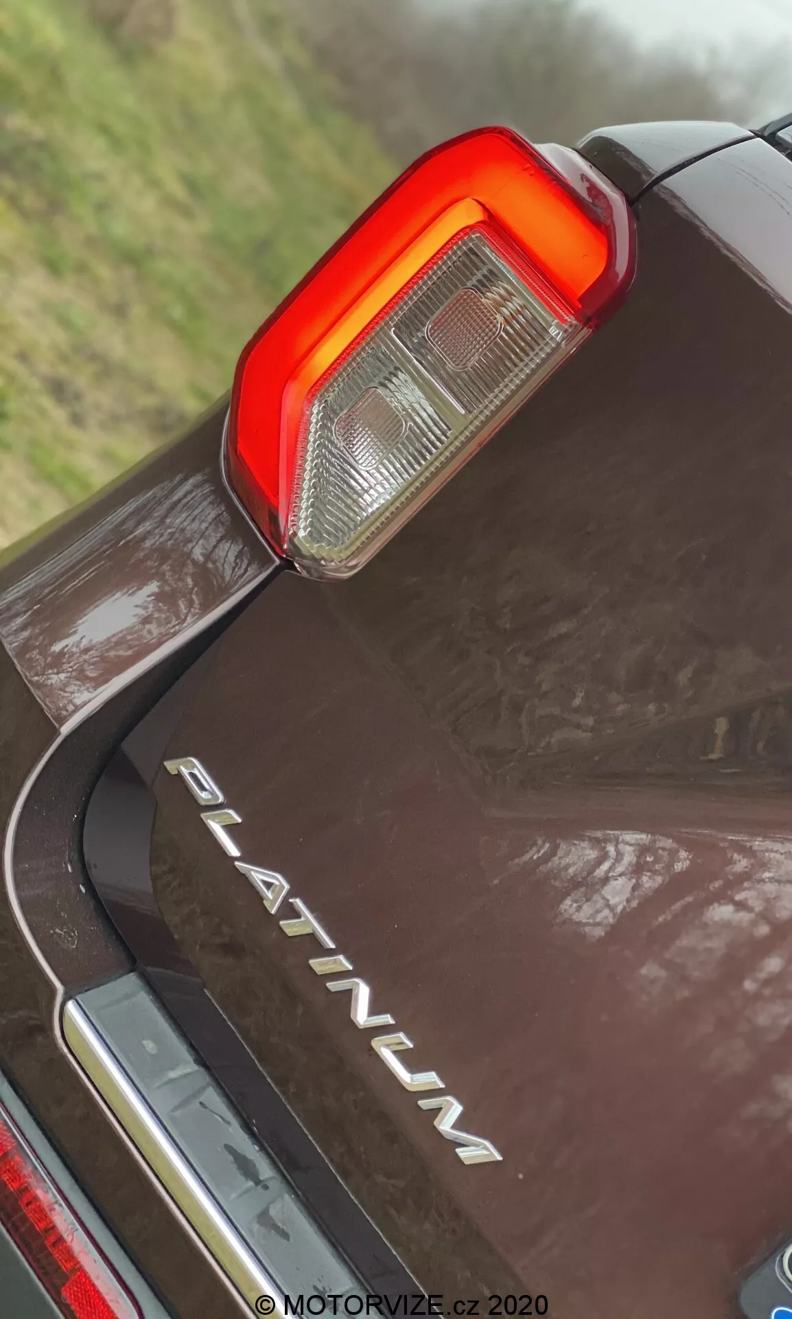 Bližnji posnetek iz nizkega zornega kota zadnjega dela Ford Explorer 2019 v različici PLATINUM, s poudarkom na navpični LED zadnji luči s prozornim in rdečim delom za vzvratne luči in zavorne luči, kromirano značko 'PLATINUM', odsevnim temnim premazom z vodnimi kapljicami in kromiranim trakom pod zadnjim odbijačem.
