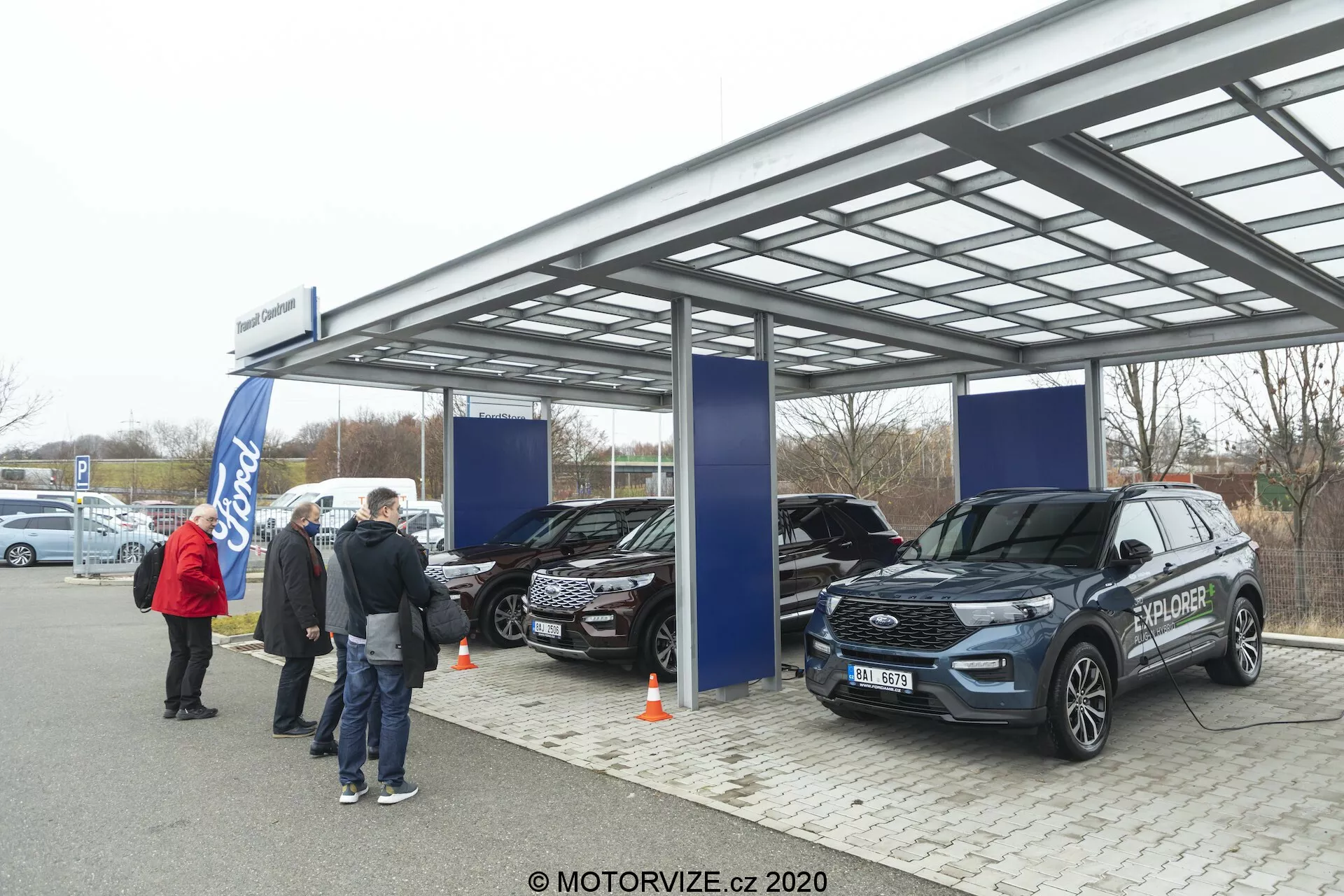 Slika prikazuje zunanji avtomobilski salon ali razstavni dogodek pod kovinskim, polprosojnim strehom z mehko, oblačno dnevno svetlobo. Trije SUV, vključno z modernim Ford Explorerjem 2019 v ospredju, so razstavljeni z vidnimi lastnostmi, kot so sprednja maska, žarometi in stranska ogledala. Ljudje in transparenti nakazujejo uradni promocijski dogodek, logotip Ford pa je viden v ozadju.