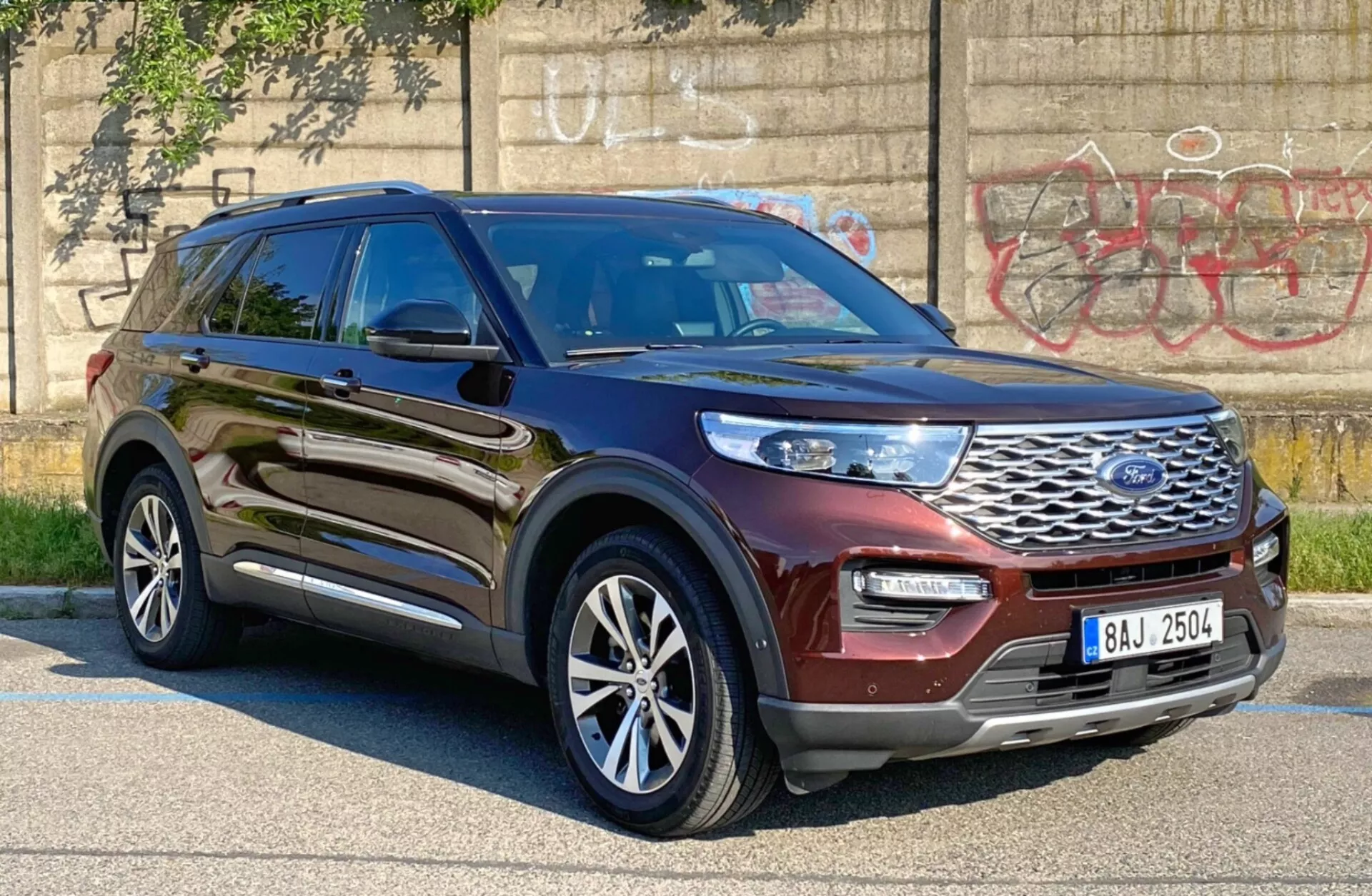 Slika SUV-ja Ford Explorer 2019, posneta pod kotom treh četrtin s prednje strani z voznikove strani, prikazuje sijočo kovinsko rjavo ali bordo barvo. Slika poudarja sprednjo rešetko z logotipom Ford, elegantne LED ali HID žaromete, sprednji odbijač in voznikov stranski profil, vključno z vrati, blatnikom in večkrakimi aluminijastimi kolesi. Vozilo je parkirano v urbanem okolju z grafiti na betonski steni v ozadju, s čimer poudarja njegovo robustno in moderno obliko, z možnimi naprednimi sistemi za pomoč vozniku, kot sta adaptivni tempomat in pomoč pri ohranjanju voznega pasu.