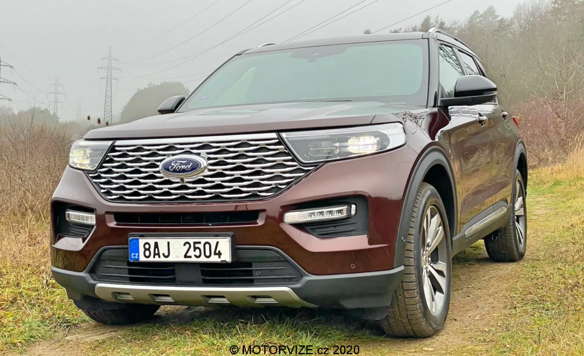 2019 Ford Explorer avtomobil fotografiran iz nizkega prednjega tridelnega kota, ki prikazuje rešetko z logotipom Ford, žaromete, meglenke, pokrov motorja, stranska okna in del zadnjega dela. Kompleksen dizajn rešetke, elegantni LED žarometi, dvobarvni odbijač in brezkontaktni sistem za vstop poudarjajo sodobne in tehnološko napredne lastnosti. Nameščen nekoliko off-road z desnimi kolesi na travi in levimi kolesi na umazani poti, kar poudarja zmožnost lahkih terenskih poti. Vidna večkraka platišča, kar kaže na celoletne pnevmatike.
