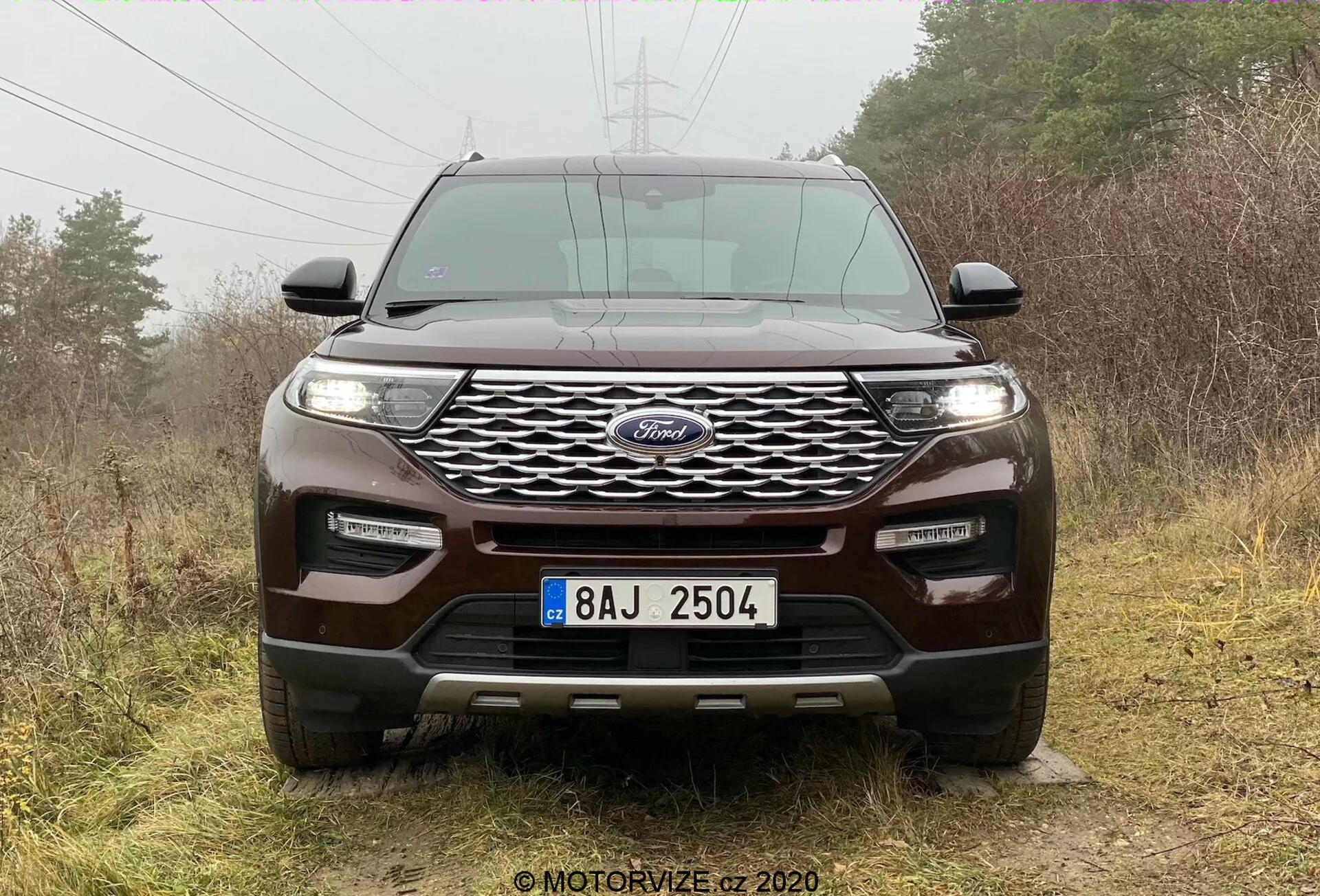 Pogled spredaj na SUV Ford Explorer 2019, ki prikazuje izrazit modri ovalni znak na sredini dinamične mrežne rešetke, obkrožen z elegantnimi vodoravno usmerjenimi žarometi, verjetno z LED ali HID lučmi. Integrirane meglenke in parkirni senzorji izboljšujejo tehnološke in varnostne značilnosti sprednjega odbijača. Kipkasta havba z dvignjenimi grebeni poudarja športno obliko, medtem ko pomemben odmik od tal, zaščitna plošča in vlečne kavlje nakazujejo terenske sposobnosti. SUV je postavljen v zunanjem, naravnem ozadju z razpršeno oblačno svetlobo, ki poudarja njegovo obliko.