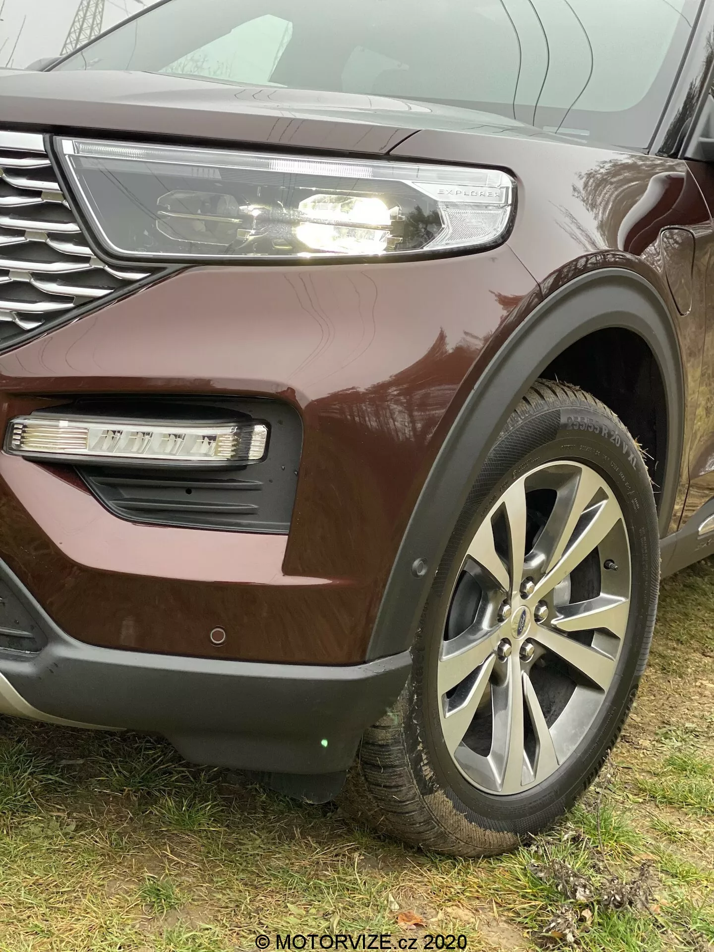 Bližnji poševni pogled na SUV Ford Explorer 2019 s poudarkom na levi sprednji četrtini, izpostavlja velika lita platišča, masko z zrcalnimi dodatki in delno vidnim LED žarometom s dnevnimi lučmi, kovinsko burgundsko barvo, meglenko v vdolbenem delu odbijača z drugačno teksturo in morebitnim vstopom za zrak ali dekorativnim elementom. Znak je na sredini večkrakih koles. Spodnji del odbijača kaže na prisotnost parkirnega senzorja ali sistema za prilagodljiv tempomat. Slika je posneta iz nizkega kota, da poudari velikost in trdnost vozila, z vidnim vodnim žigom 'MOTORVIZE.cz 2020' v spodnjem desnem kotu.