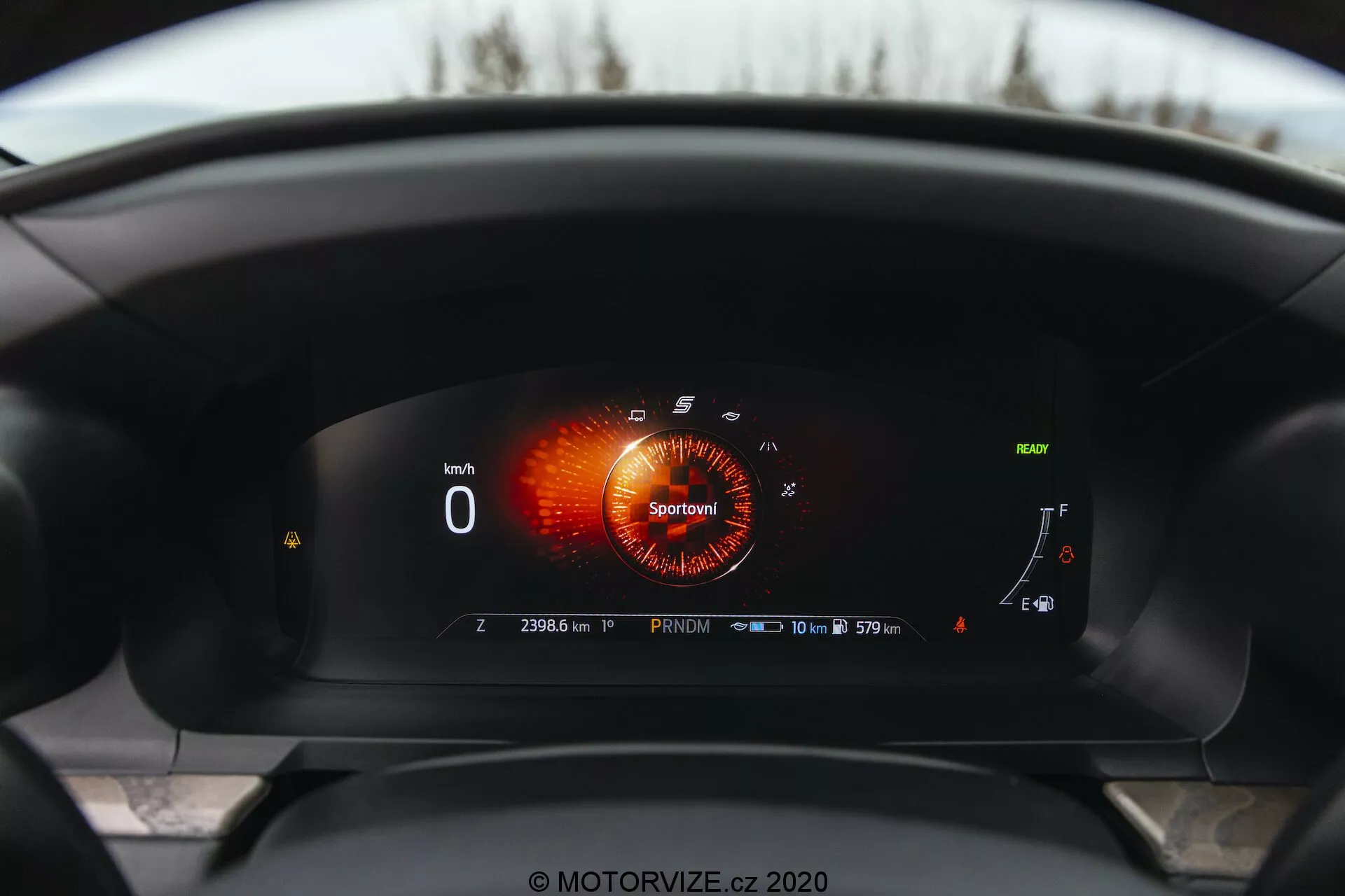 Pogled v notranjost Forda Explorer 2019 iz vozniške perspektive, prikazuje popolnoma digitalno instrumentno ploščo. Digitalni zaslon prikazuje merilnik vrtljajev v načinu 'Sport' z rdeče-oranžno grafiko, merilnik hitrosti, ki kaže '0 km/h,' skoraj poln merilnik goriva, merilnik temperature hladilne tekočine na spodnjem koncu in različne indikatorje. Opažena je tudi simbol adaptivnega tempomata, opomnik na servisni interval, ki kaže '10 000 km ali 365 dni,' in števec kilometrov na '23063 km,' kar kaže, da je vozilo ustavljeno v načinu 'Park.' Ikona skoraj polne baterije nakazuje hibridno ali električno zmogljivost.