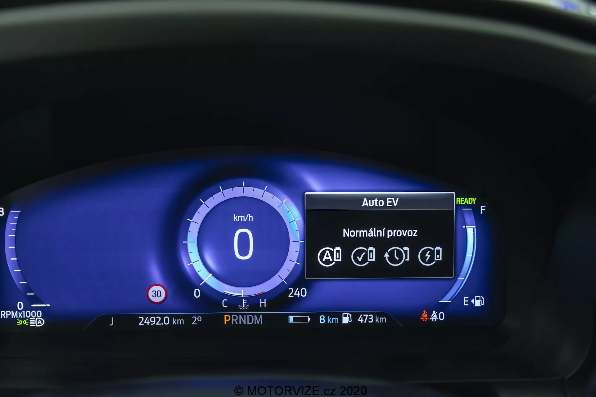 Prikaz od blizu digitalne instrumentne plošče Ford Explorer 2019 iz vozniškega zornega kota. Digitalni merilnik hitrosti kaže 0 km/h in instrumentna plošča vključuje način 'Auto EV', ki nakazuje električni ali hibridni pogonski sistem, stanje 'Ready', ikone za funkcije samodejne vožnje, števec kilometrov na 24.920 km, indikator prestav PRNDM, ikone za stanje za baterijo in zunanje osvetlitev, prikaz porabe goriva z 8 km preostanka in največjim dosegom 473 km, digitalni merilnik goriva in brez opozorilnih lučk. Modra barvna shema ponuja sodobno estetiko.