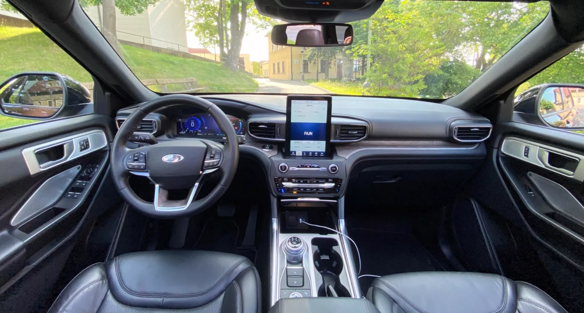 Pogled v notranjost Ford Explorer 2019 iz perspektive zadnjih sedežev, obrnjeno proti armaturni plošči. Slika zajema simetrično postavitev, vključno z volanom z večfunkcijskimi gumbi, zaslonom na dotik infotainment sistema, konzolo menjalnika z nadzornimi gumbi in oblazinjenjem sedežev ter vratnih panelov, ki posnema usnje. Notranjost vrhunskega razreda je poudarjena z elegantnimi kovinskimi dodatki in dobro nameščeno osvetlitvijo.