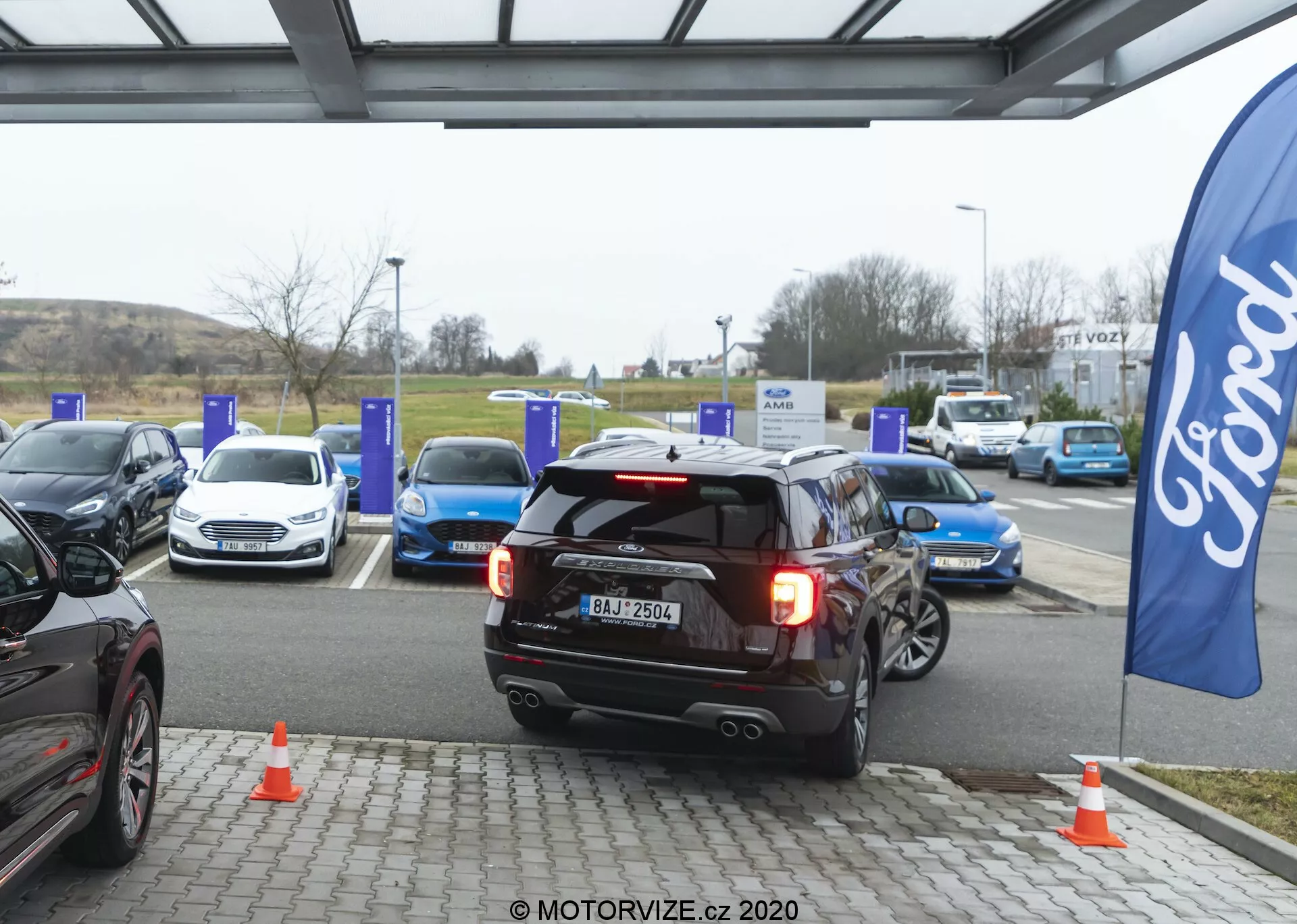 Črni SUV Ford Explorer 2019, postavljen v sredino, bodisi vzvratno parkira ali se oddaljuje od EV polnilnega mesta na polnilni postaji. Zadnje luči so prižgane, kar lahko nakazuje na vzvratno vožnjo ali zaviranje, z vidno registrsko tablico, dvojnim športnim izpušnim sistemom, zadnjim brisalcem in visoko nameščeno zadnjo zavorno lučjo. Okolica prikazuje več električnih vozil pri modrih polnilnih stebrih in oranžne prometne stožce blizu SUV-ja. Zastava z logotipom Ford nakazuje prisotnost znamke. Prizor je posnet v oblačnem dnevu z difuzno naravno svetlobo, kar poudarja avtomobilsko tehnologijo in trajnostno prometno infrastrukturo.