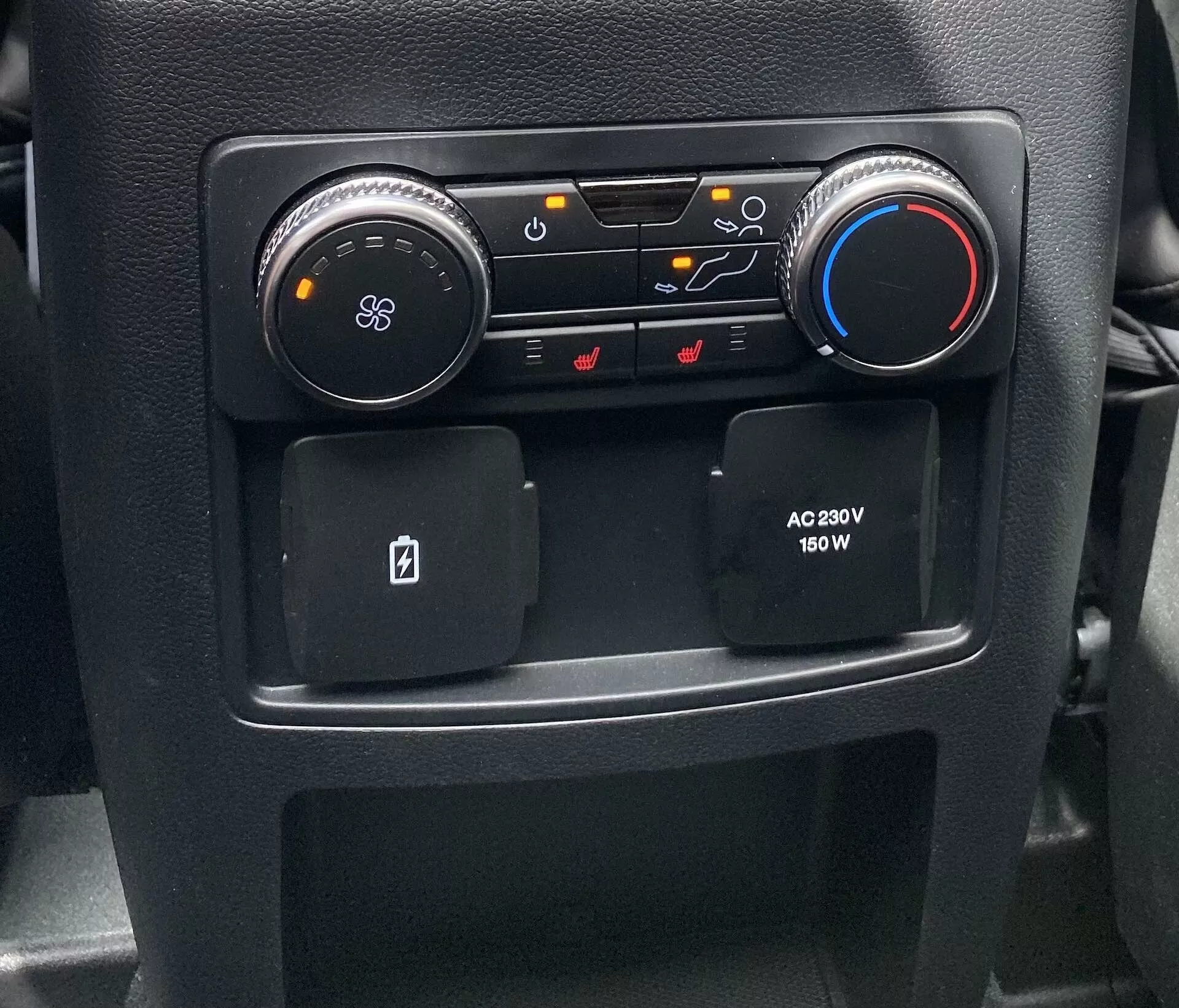 Pogled od blizu na del osrednje konzole notranjosti avtomobila Ford Explorer 2019, ki prikazuje nadzor HVAC in udobja. Na levi strani je okrogel črn gumb z ikono ventilatorja za nastavitev zračenja; poleg njega so gumbi za usmerjanje pretoka zraka, kroženje zraka in funkcije za odmrzovanje vetrobranskega stekla. Na desni strani je modro-rdeč gumb za nastavitev temperature. Spodaj sta dva protizdrsna predalčka za shranjevanje, eden je namenjen kartici, ter omarica za napajanje na strani potnika z močjo 230V in 150W za elektronske naprave.