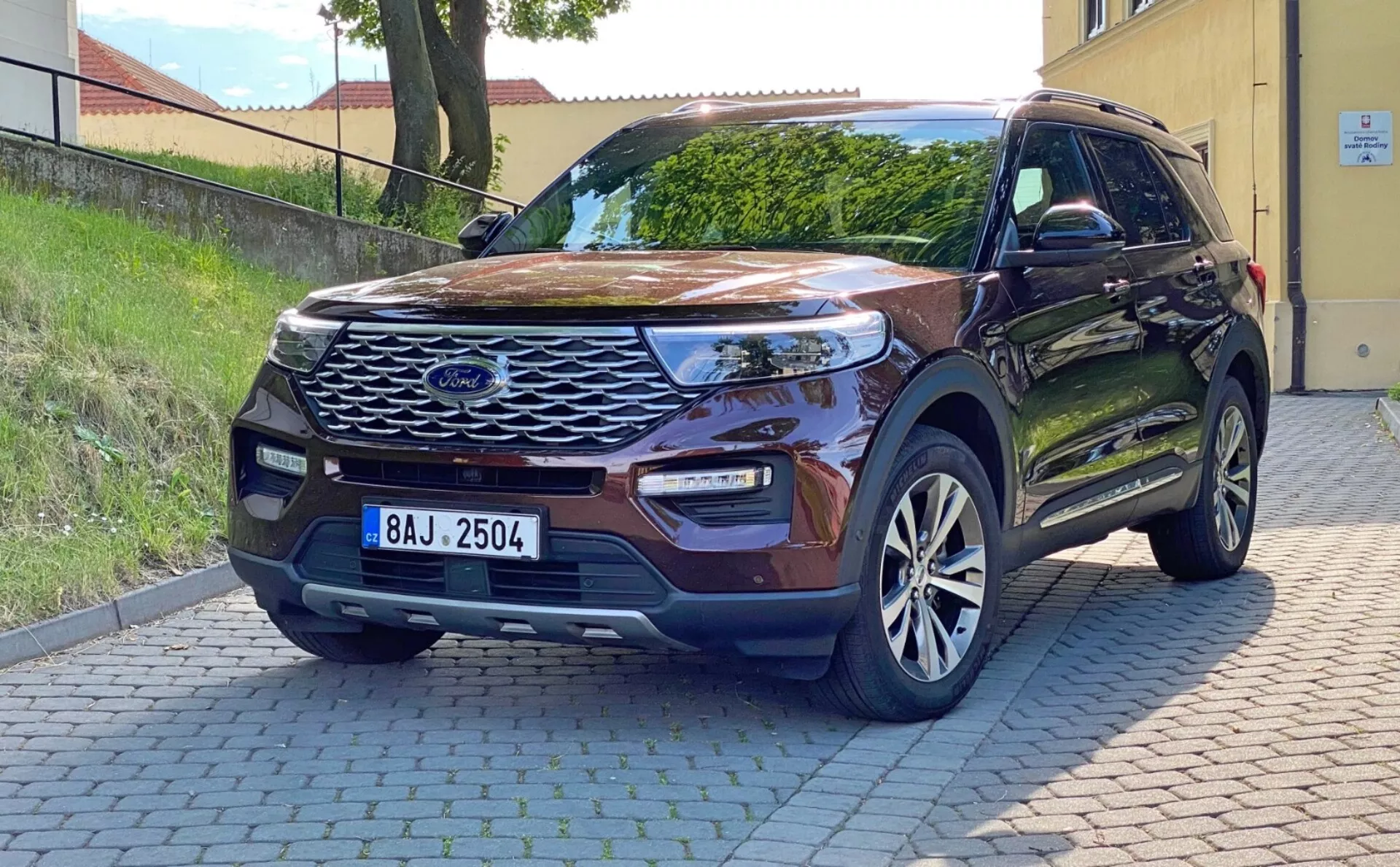 Slika sodobnega SUV Ford Explorer 2019 parkiranega na asfaltirani površini, posneta iz sprednje trikotne perspektive. Prikazuje izrazito sprednjo masko s sredinskim emblemom, elegantne LED žaromete, meglenke vgrajene v odbijač, parkirne senzorje, izklesano pokrov motorja, aluminijasta platišča, stranska ogledala v barvi vozila z vgrajenimi smerniki, brezključni vstop in stanovanjsko ozadje. Naravna dnevna svetloba poudarja njegove zunanje lastnosti in tehnološke izboljšave.