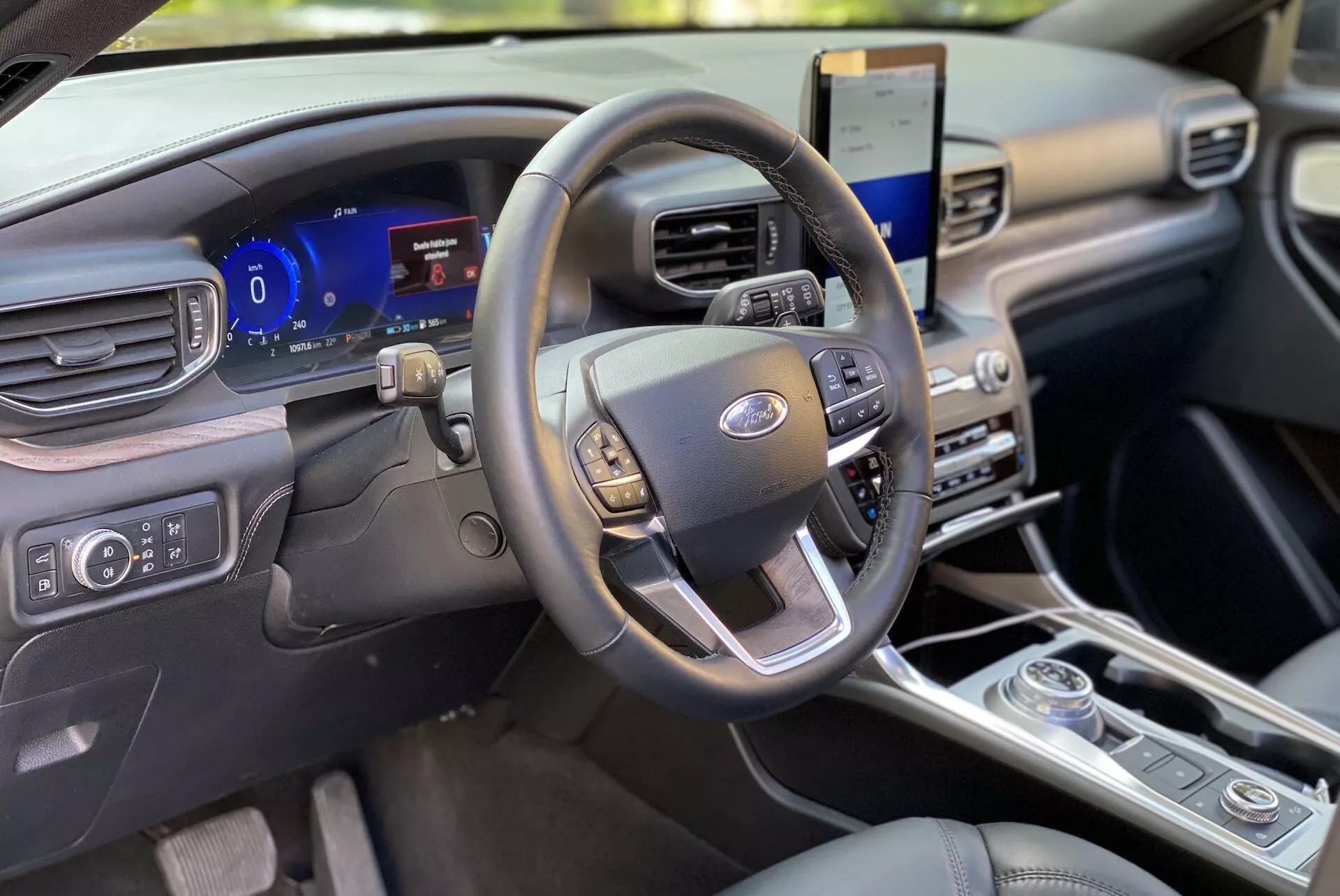 Notranjost Forda Explorer 2019, s poudarkom na območju voznika iz perspektive sprednjega potnika. Volan, z večfunkcijskimi kontrolami za avdio, tempomat in brezstično komunikacijo, je centriran. Za njim je digitalni instrumentni grozd, ki prikazuje prilagodljive merilnike in podatke vozila v realnem času. Na desni strani je navpično usmerjen zaslon na dotik, ki prikazuje informacijski sistem z satelitsko navigacijo in integracijo pametnih telefonov. Spodaj so večcona klimatska kontrola in vrtljiv izbirnik prestav, kar kaže na sodoben dizajn. Plošča kabine je čista in moderna, narejena iz premium materialov, oddaja minimalistično estetiko.
