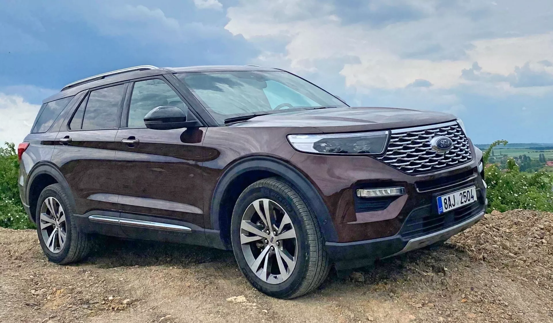 Slika SUV vozila Ford Explorer 2019, parkiranega na nagnjeni, morda neasfaltirani površini z temnim odtenkom in blatom poškropljenimi stranicami, izrazito sprednjo rešetko z mrežasto zasnovo, LED žarometi, meglenkami, aluminijastimi platišči z večkrakim dizajnom, stranskimi ogledali z integriranimi smerniki, strešnimi nosilci, stransko stopnico, rahlo zatemnjenimi okni in anteno na zadnjem delu strehe, ki poudarjajo njegove robustne zmogljivosti in razkošne lastnosti v podeželskem ali terenskem okolju.