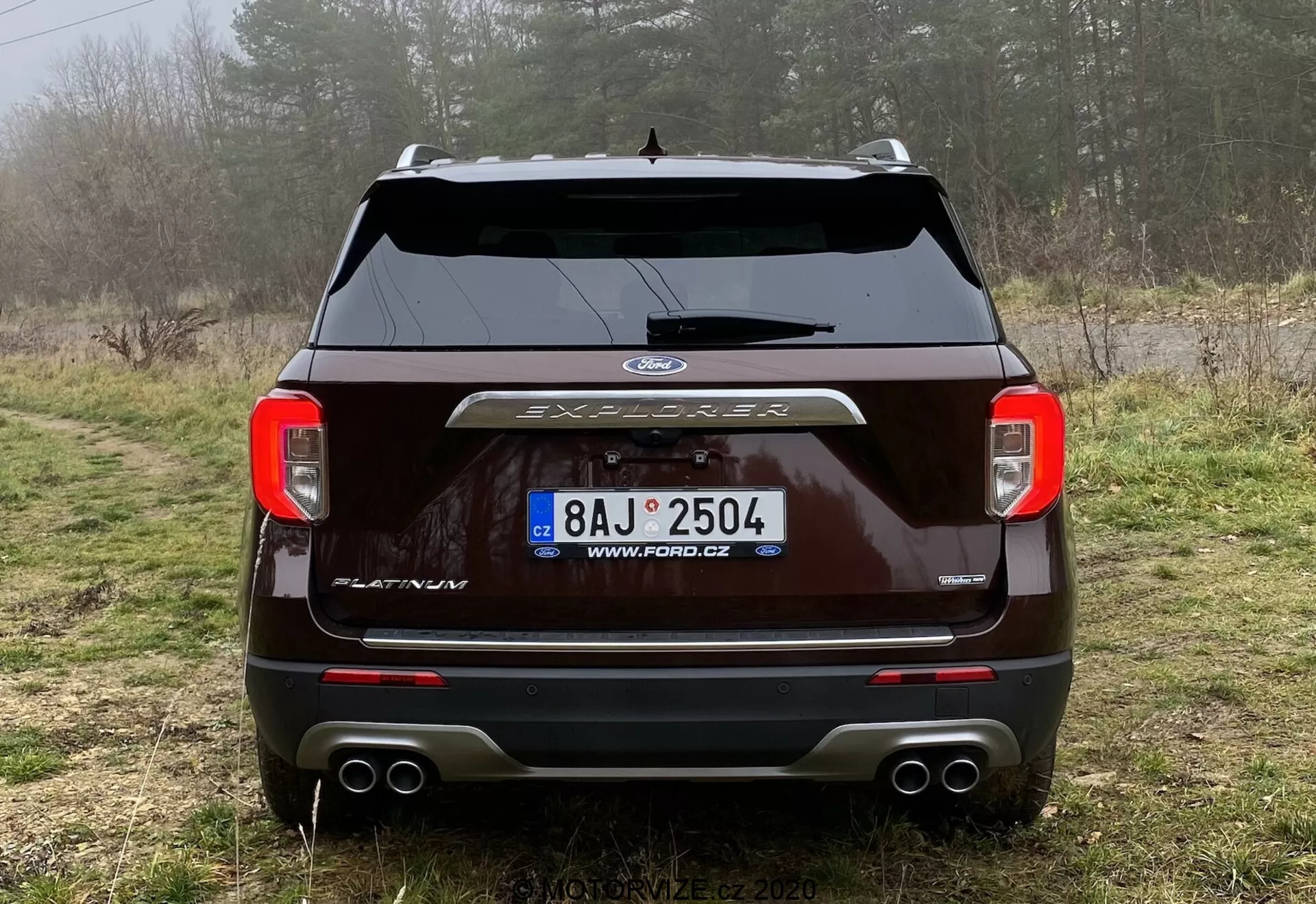Zadnji pogled na temno obarvan Ford Explorer 2019, parkiran na makadamski poti v podeželskem okolju z vegetacijo in meglo. Slika prikazuje jasne zadnje luči, centralno nameščen logotip Ford, napis 'EXPLORER', zadnji brisalec, parkirne senzorje, dvojni izpušni sistem in pokrov vlečne kljuke. Videti je registrsko tablico s češko URL, ki poudarja zunanjo obliko in tehnologijo vozila.